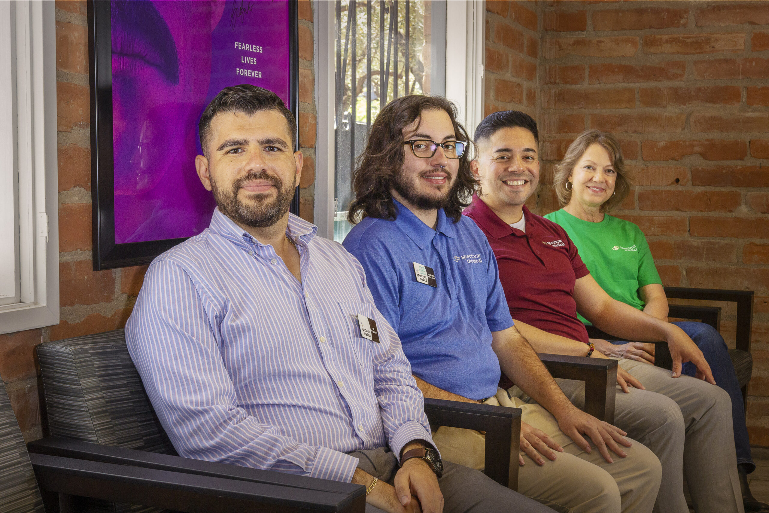 Left to right Dr Jose Rodriguez Garcia, Ari Kravitz, Victor Avila, Alecia Alfredson: A group of four individuals sits in a cozy office space with a brick wall and large windows in the background. They are dressed in various professional and branded shirts, smiling warmly at the camera. A bright purple poster with the words "Fearless Lives Forever" is prominently displayed on the wall, adding a motivational touch to the setting.