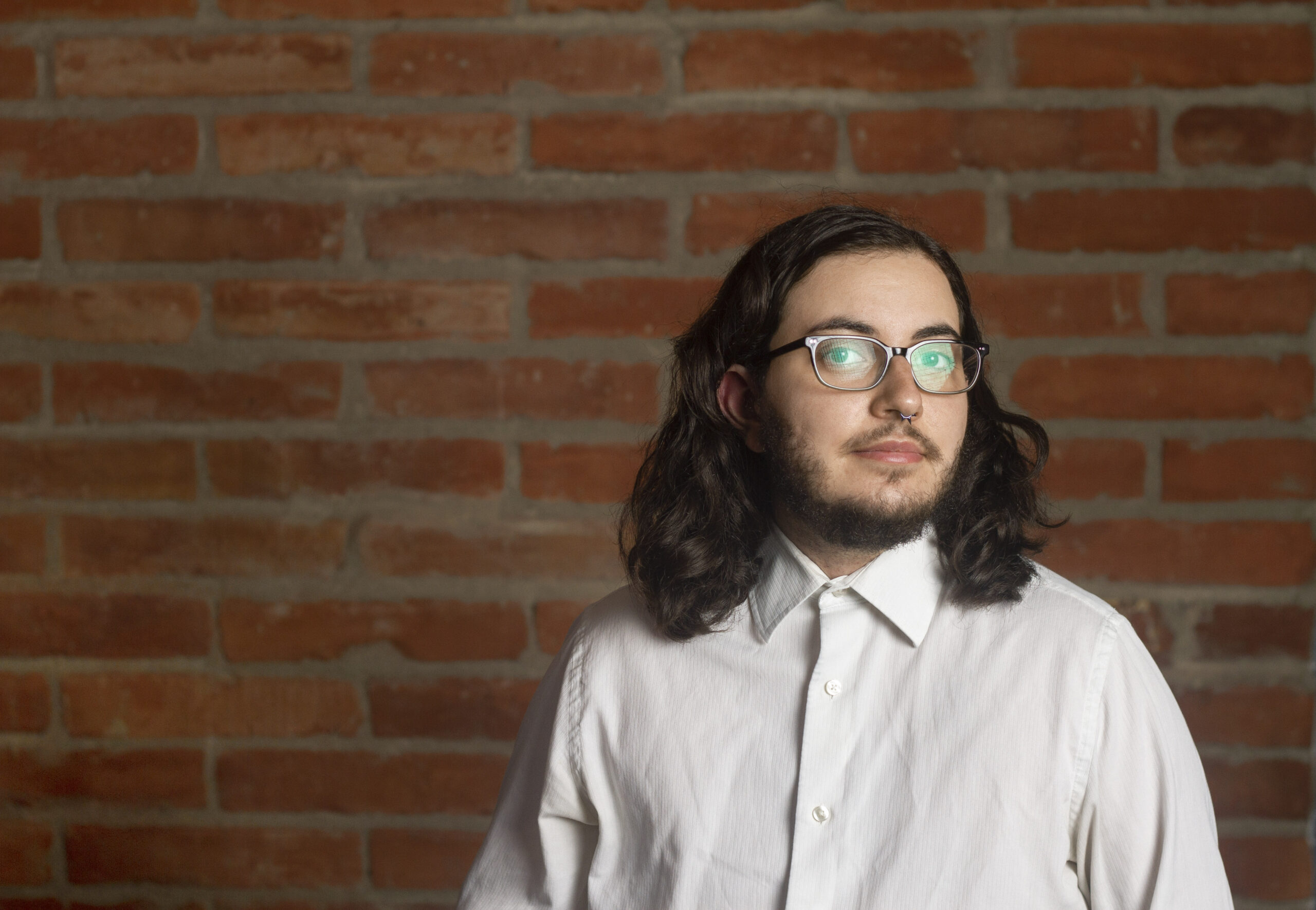 Ari Kravitz. A portrait of a person with long dark hair, glasses, and a nose ring, wearing a crisp white shirt. They are standing against a red brick wall, with soft lighting creating a warm and thoughtful atmosphere.