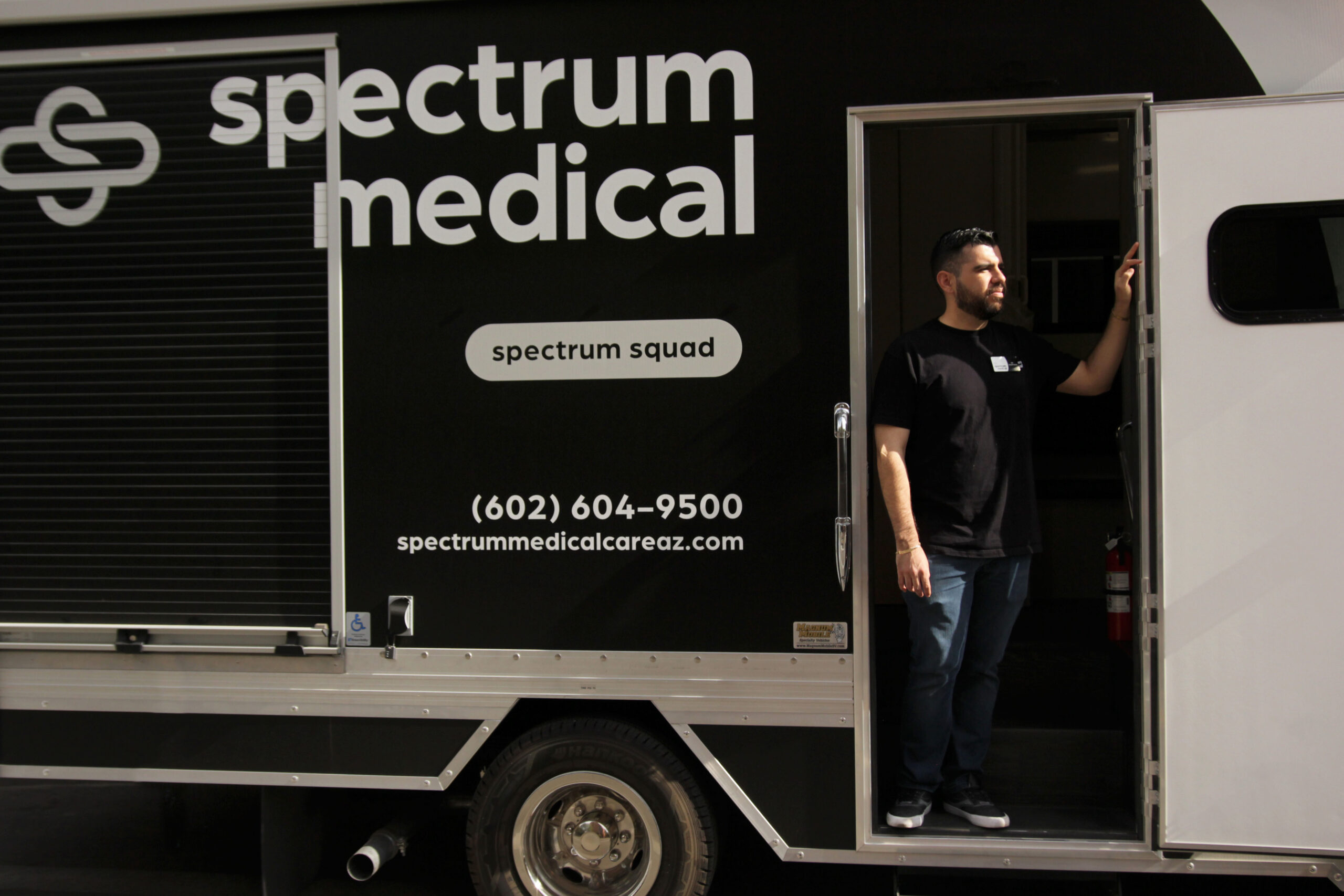 A man stands at the open door of a "Spectrum Medical" mobile unit, wearing casual jeans and a black T-shirt. The vehicle prominently displays the "Spectrum Medical" logo along with the text "Spectrum Squad" and contact information, including a phone number and website. The scene highlights mobile healthcare services in an accessible and professional manner.
