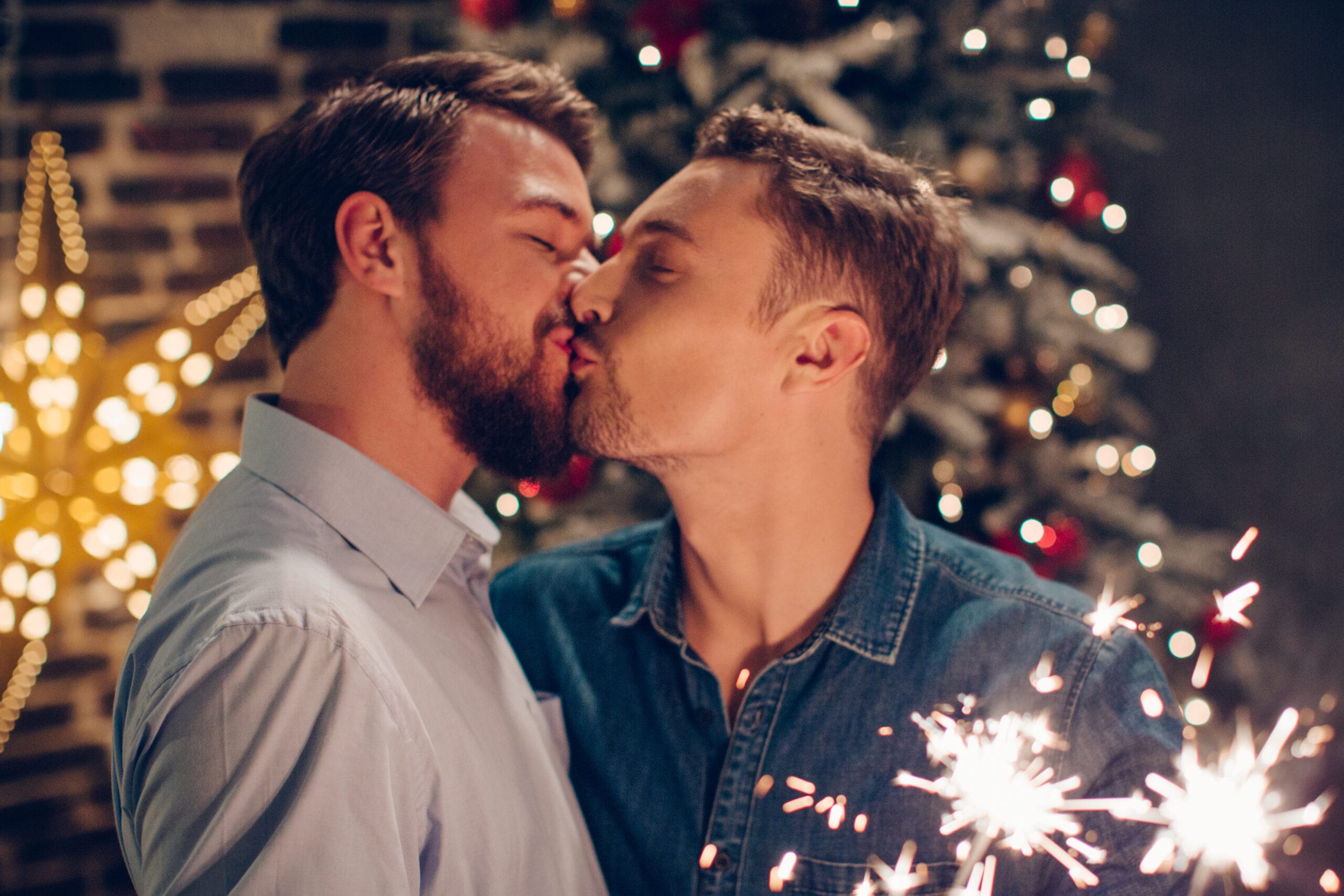 Two men share a kiss in a warmly lit holiday setting, with sparklers glowing in their hands. They stand in front of a decorated Christmas tree with twinkling lights and ornaments, as well as a star-shaped festive decoration in the background, creating a romantic and celebratory atmosphere.