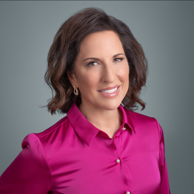 Amy Sneor, RN, BSN,BA(She/Her), A professional portrait of a smiling woman with short, wavy brown hair, wearing a vibrant magenta blouse with pearl accents and gold earrings. She is posed against a soft gray background, exuding a confident and approachable demeanor.