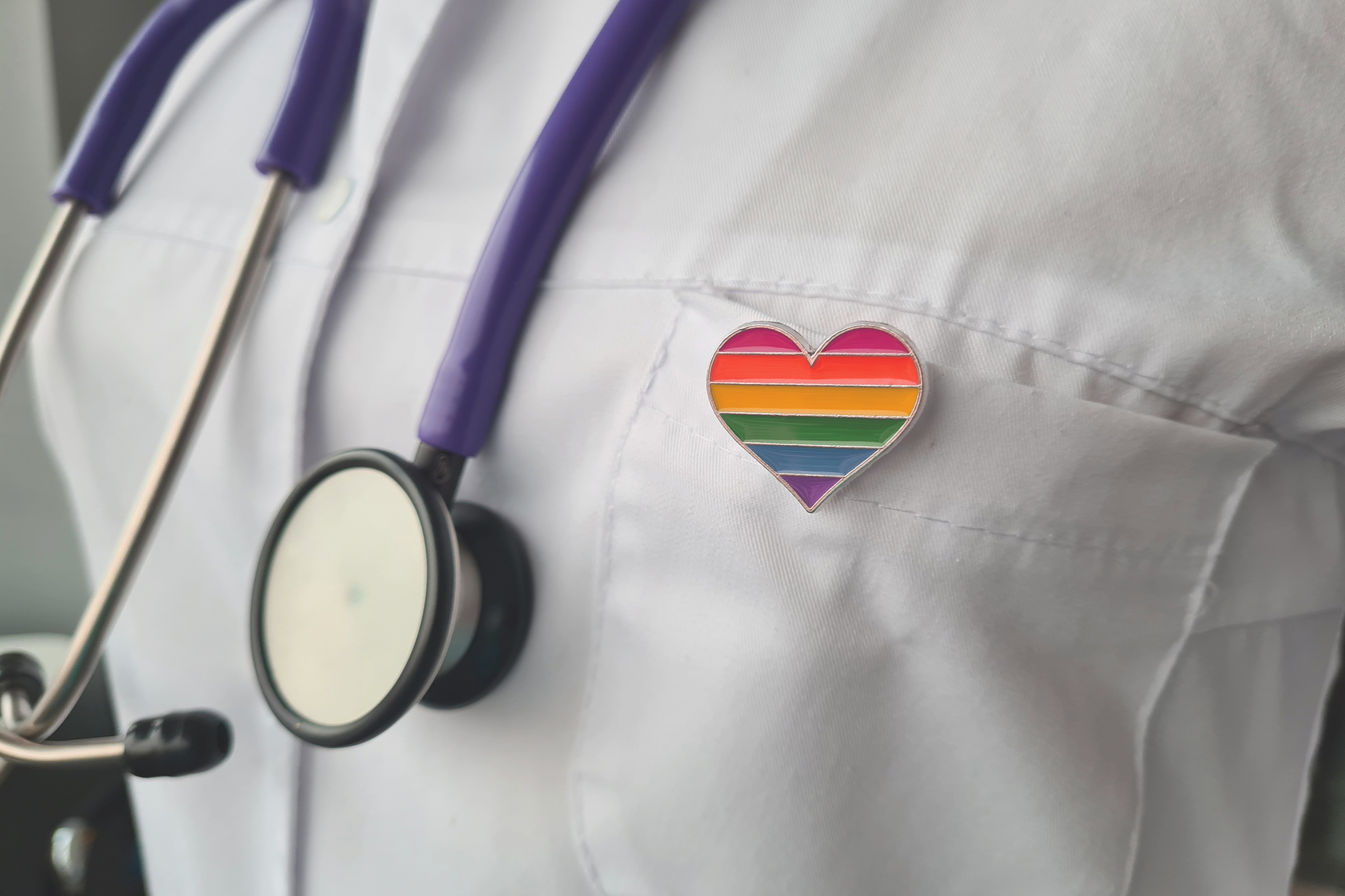 A close-up image of a healthcare professional's white coat with a purple stethoscope draped around their neck and a heart-shaped pin featuring the rainbow pride flag on the chest pocket.