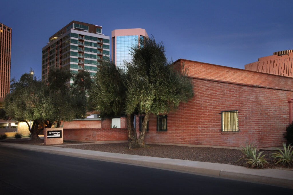 A red-brick building with a sign reading'Spectrum Medical' in front, surrounded by trees and a landscaped area. In the background, modern high-rise buildings and a deep twilight sky create a contrast between the traditional and contemporary architecture.