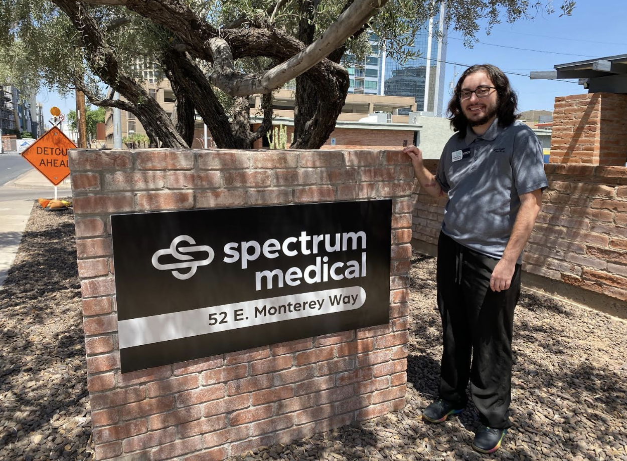 A person standing next to a brick sign for Spectrum Medical at 52 E. Monterey Way, surrounded by trees and urban buildings in the background.