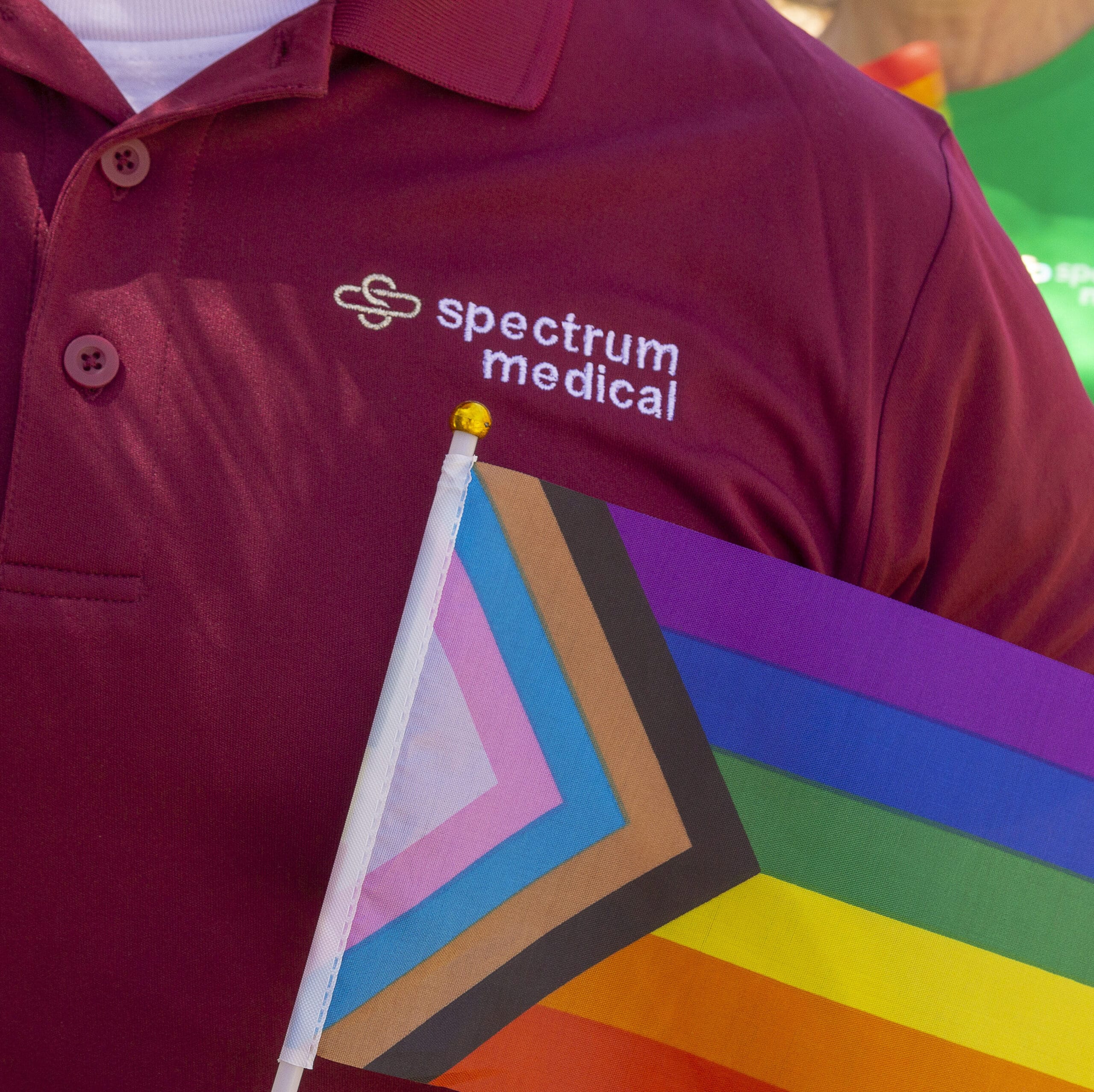 A close-up image of a person wearing a maroon polo shirt with the "Spectrum Medical" logo embroidered on it, holding a Progress Pride flag with vibrant colors and a gold-tipped flagpole.