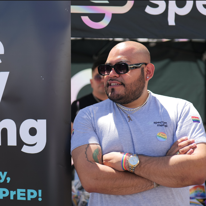 A person wearing a light gray "Spectrum Medical" t-shirt smiles while standing outdoors under a branded canopy. They sport sunglasses, a watch, colorful bracelets, and a pin reading "I am loved," radiating confidence and positivity. The setting appears to be a community event promoting inclusivity and health awareness, as suggested by the partial text visible on a nearby banner.