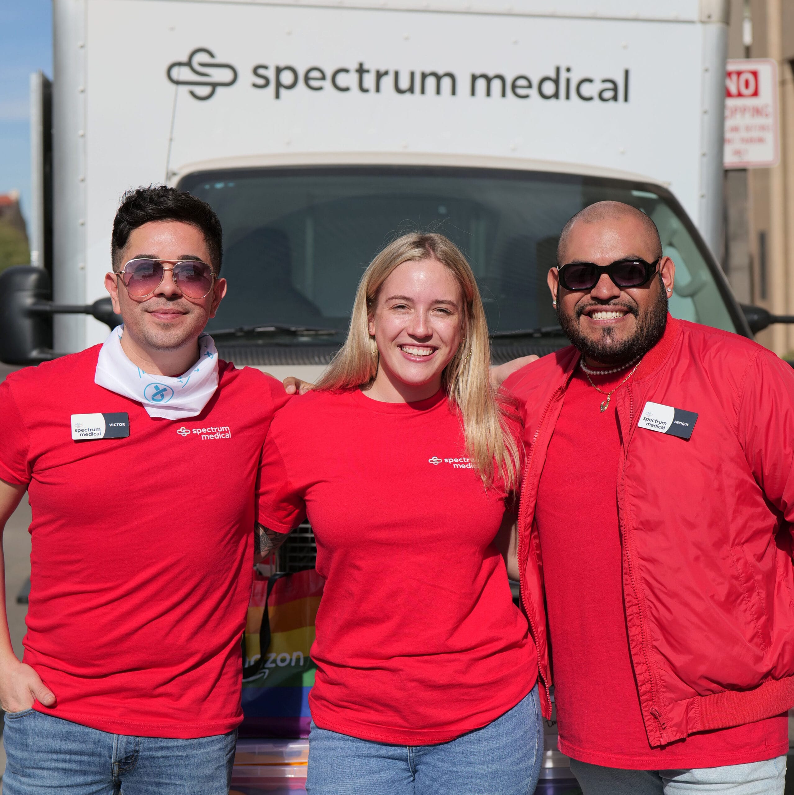 Three smiling individuals wearing matching red "Spectrum Medical" t-shirts stand in front of a Spectrum Medical van. The group exudes positivity, with the central figure, a blonde woman, flanked by two men—one wearing sunglasses and a red jacket, the other sporting a white bandana. A rainbow flag is partially visible below the van's windshield, suggesting an inclusive and welcoming event.