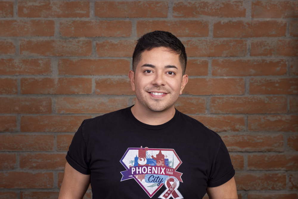 Victor Avila, smiling stands in front of a textured brick wall, wearing a black t-shirt with a graphic that reads "Phoenix City" and features city imagery and a ribbon design. The warm lighting emphasizes their friendly expression and the casual yet polished vibe of the portrait.