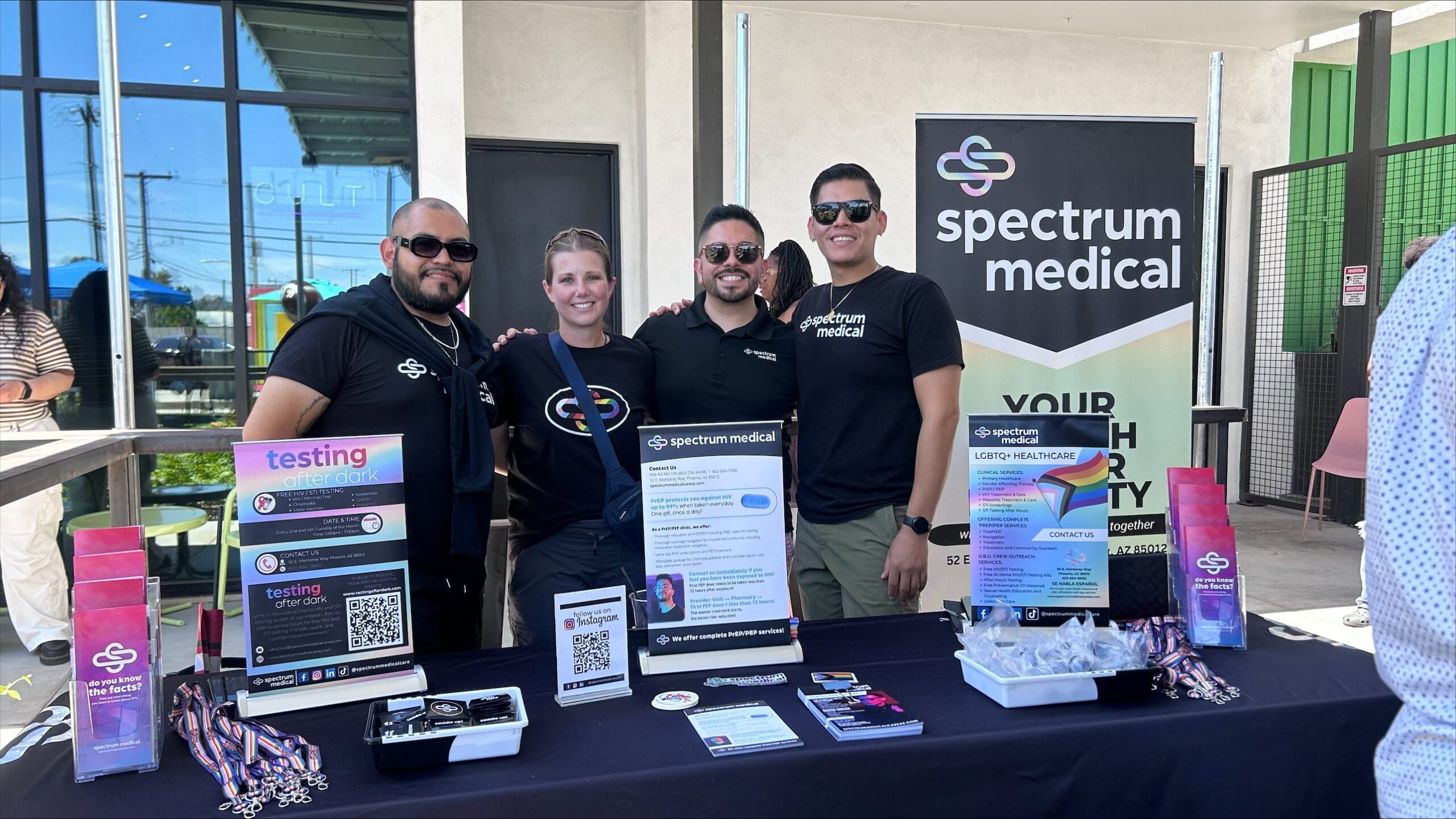 A group of four individuals stands behind a Spectrum Medical booth at an outdoor event, smiling for the camera. The booth features colorful signage about LGBTQIA2S+ healthcare services, PrEP/PEP, and free STI testing, with informational flyers, keychains, and brochures displayed on the table. A banner in the background reads "Spectrum Medical," emphasizing inclusive healthcare.