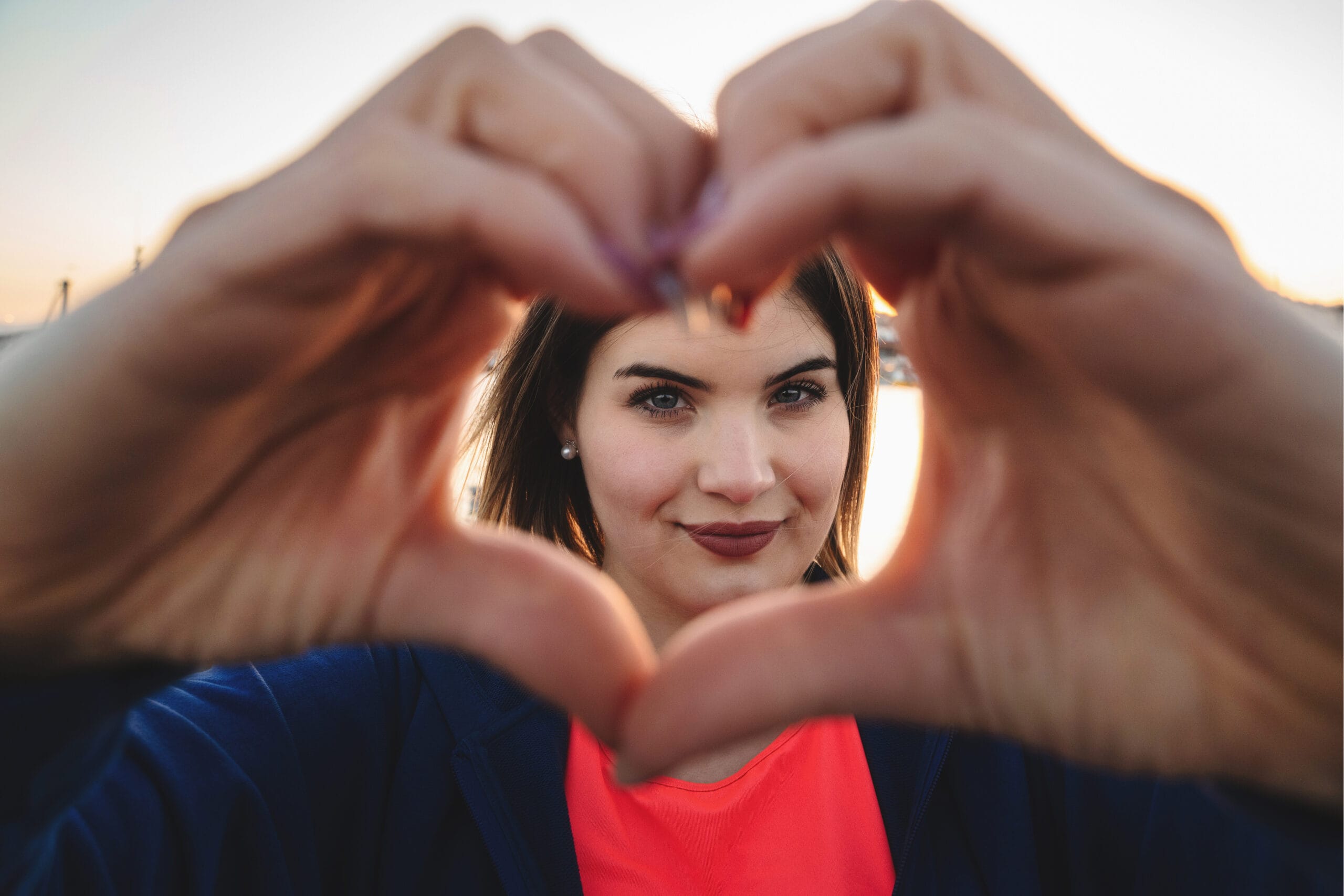 A woman framed through her hands forming a heart shape, with a serene outdoor background softly lit by a setting sun. She has a confident smile, wears a bright pink top under a blue jacket, and exudes warmth and positivity. The focus emphasizes the heartfelt gesture and her expressive gaze.