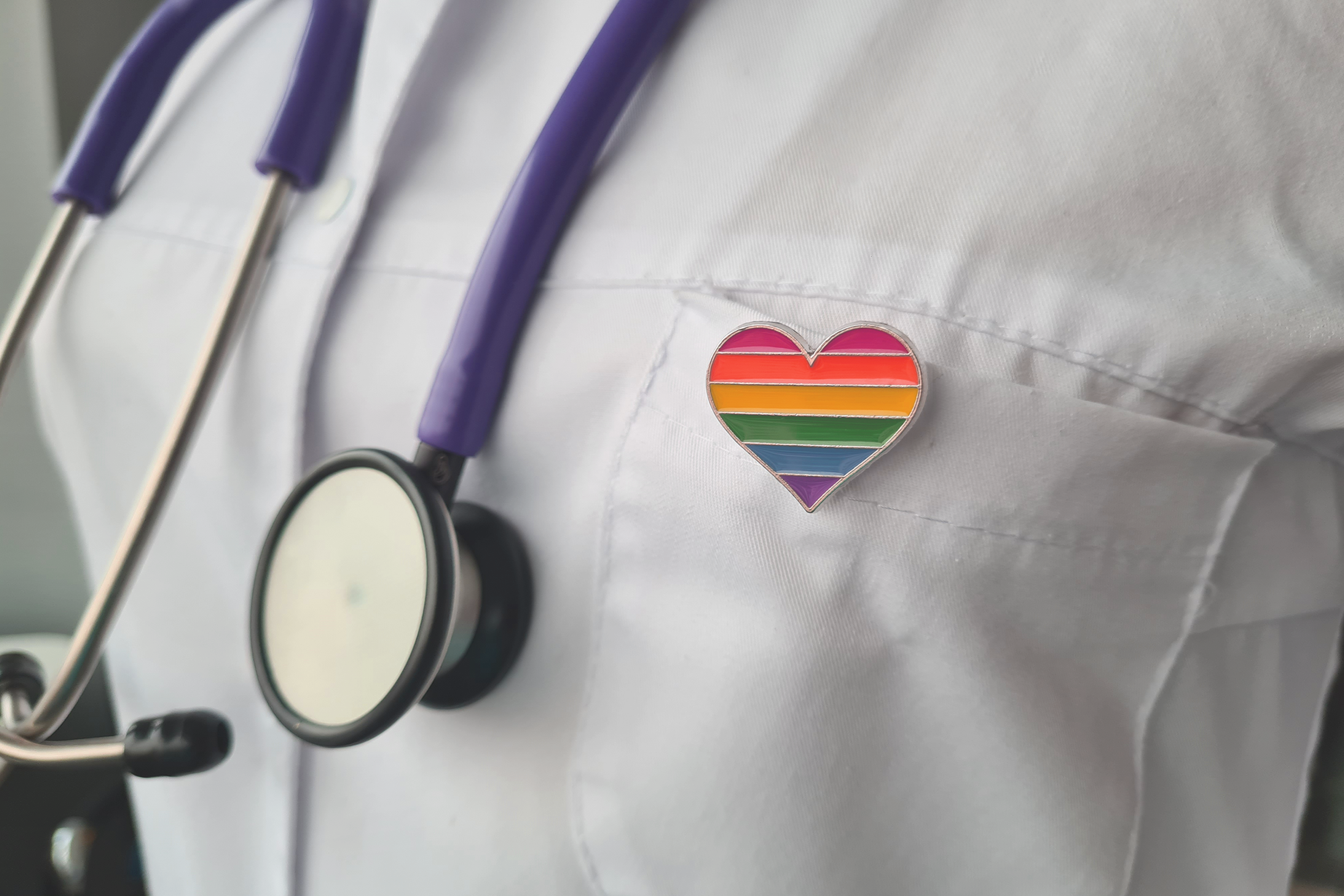 A stethoscope and a rainbow heart pin on a white lab coat, symbolizing support for LGBTQIA2S+ healthcare.