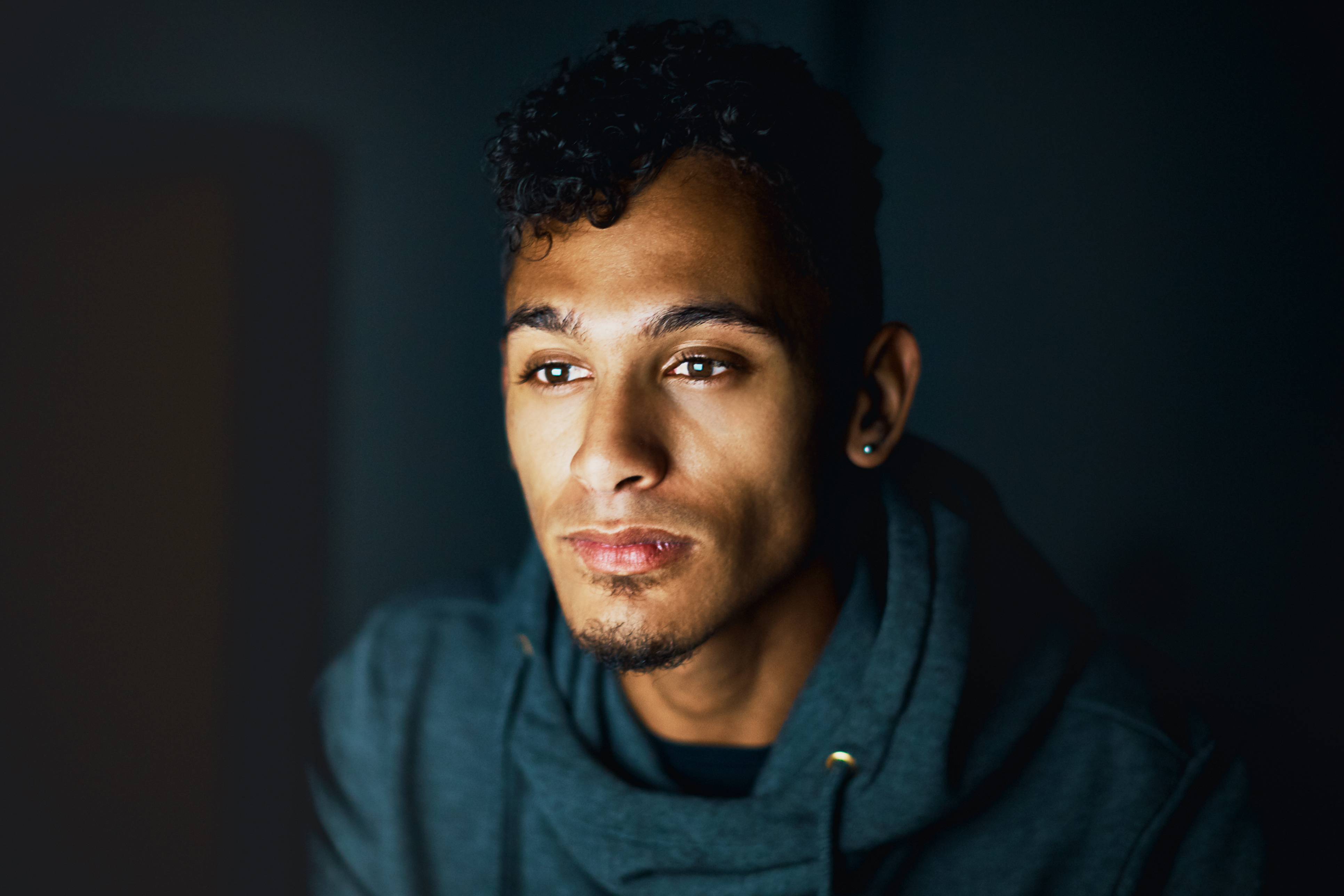 A contemplative person with short, curly hair and a small goatee gazes thoughtfully off-camera in a dimly lit setting. They are wearing a dark hoodie, and their face is softly illuminated, highlighting their focused expression. The moody lighting adds a sense of introspection and depth to the portrait.