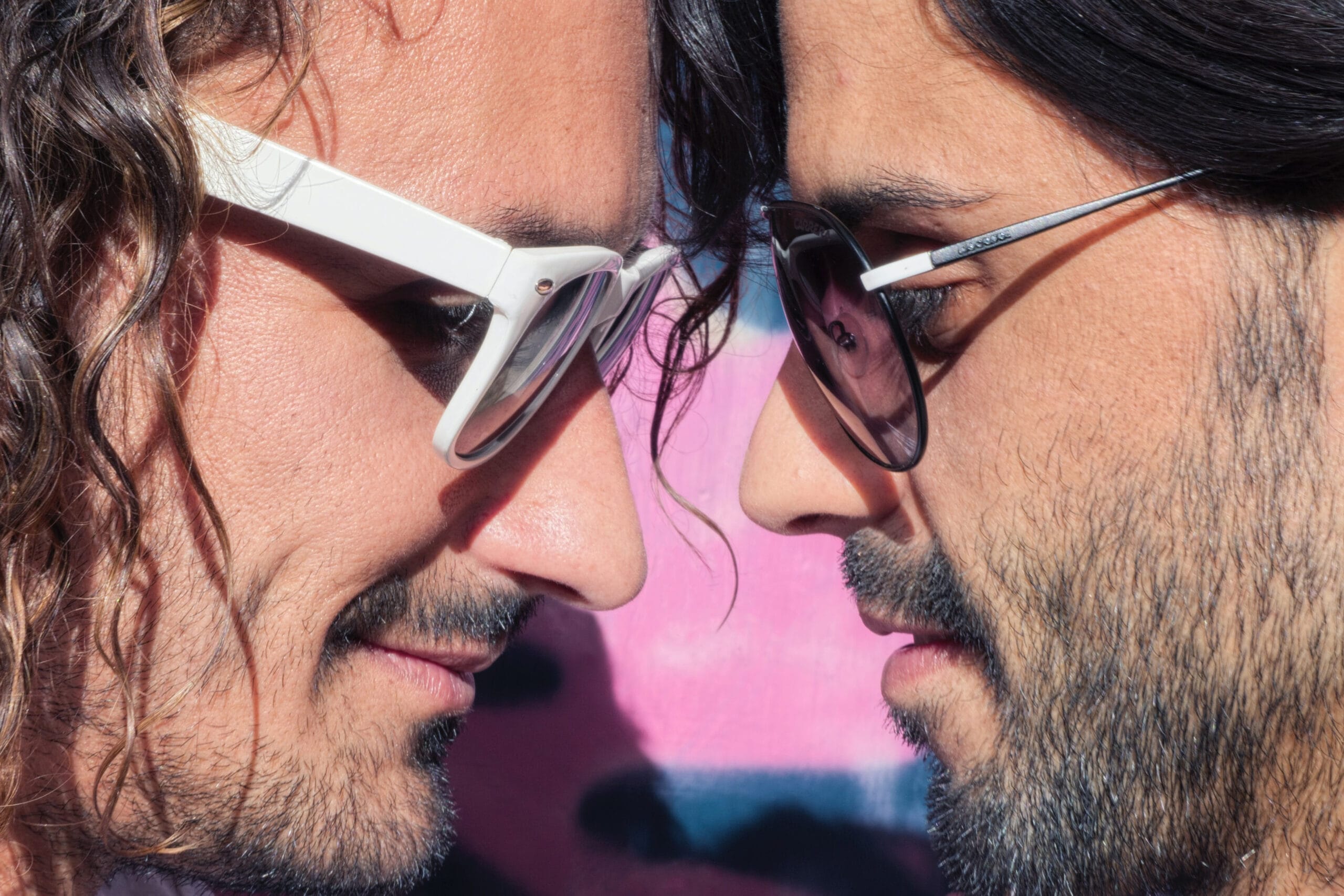 A close-up shot of two individuals wearing sunglasses facing each other, with their noses nearly touching. One person sports white-framed glasses and wavy hair, while the other has dark-framed sunglasses and a neatly trimmed beard. The background features vibrant pink tones, enhancing the intimate and stylish mood of the image.