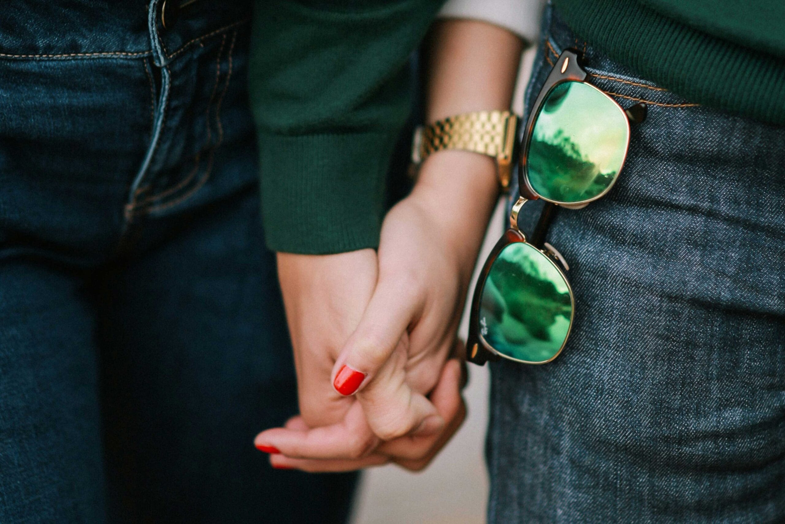 Two people holding hands, with a pair of sunglasses tucked into the back pocket of one person's jeans. This image represents love, companionship, or a shared experience.