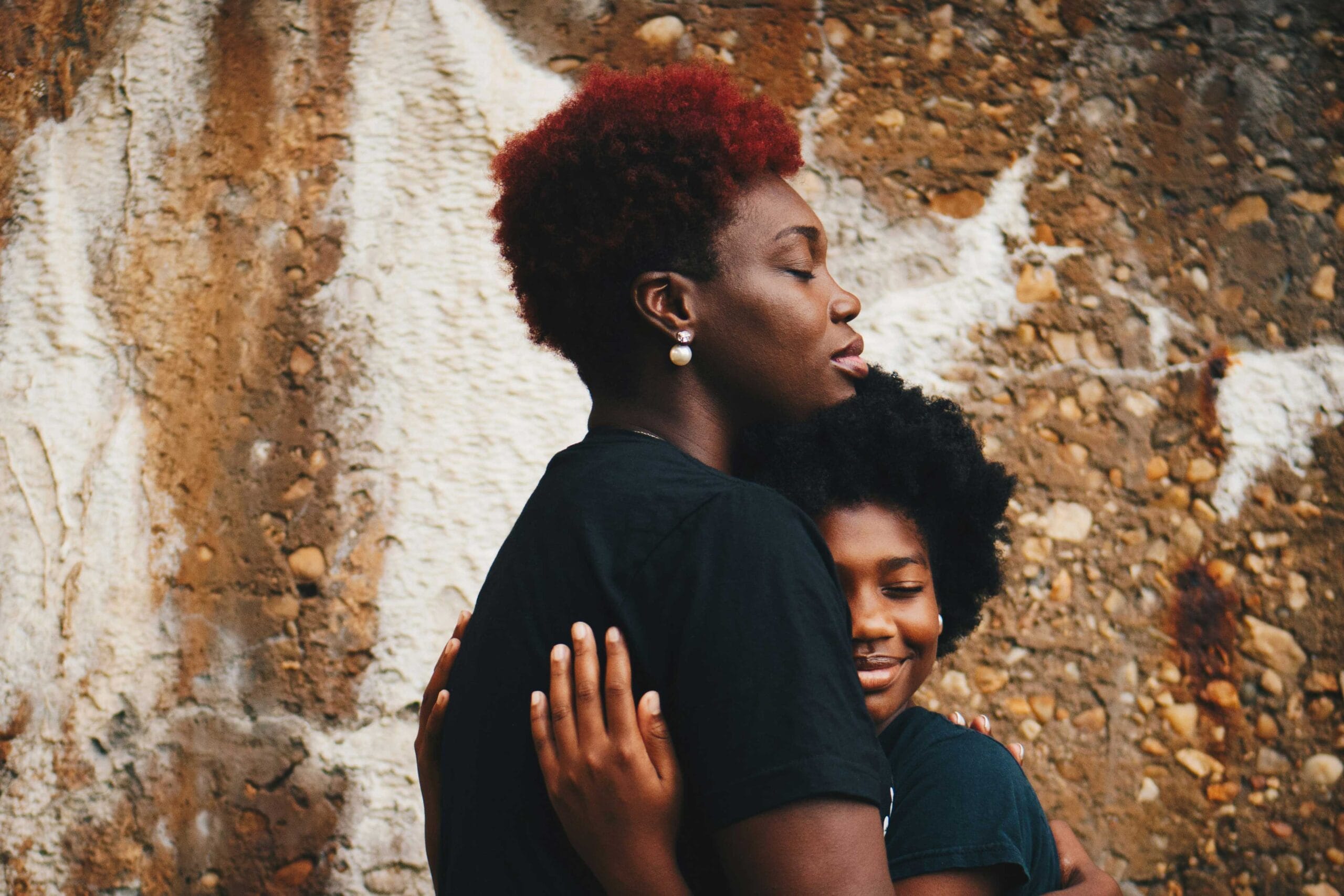 A heartfelt moment of a woman with vibrant red hair embracing a child with a warm smile and natural black curls. Both are dressed in black against a textured stone wall backdrop, exuding love, comfort, and connection. The woman's eyes are closed, capturing a sense of peace and serenity.