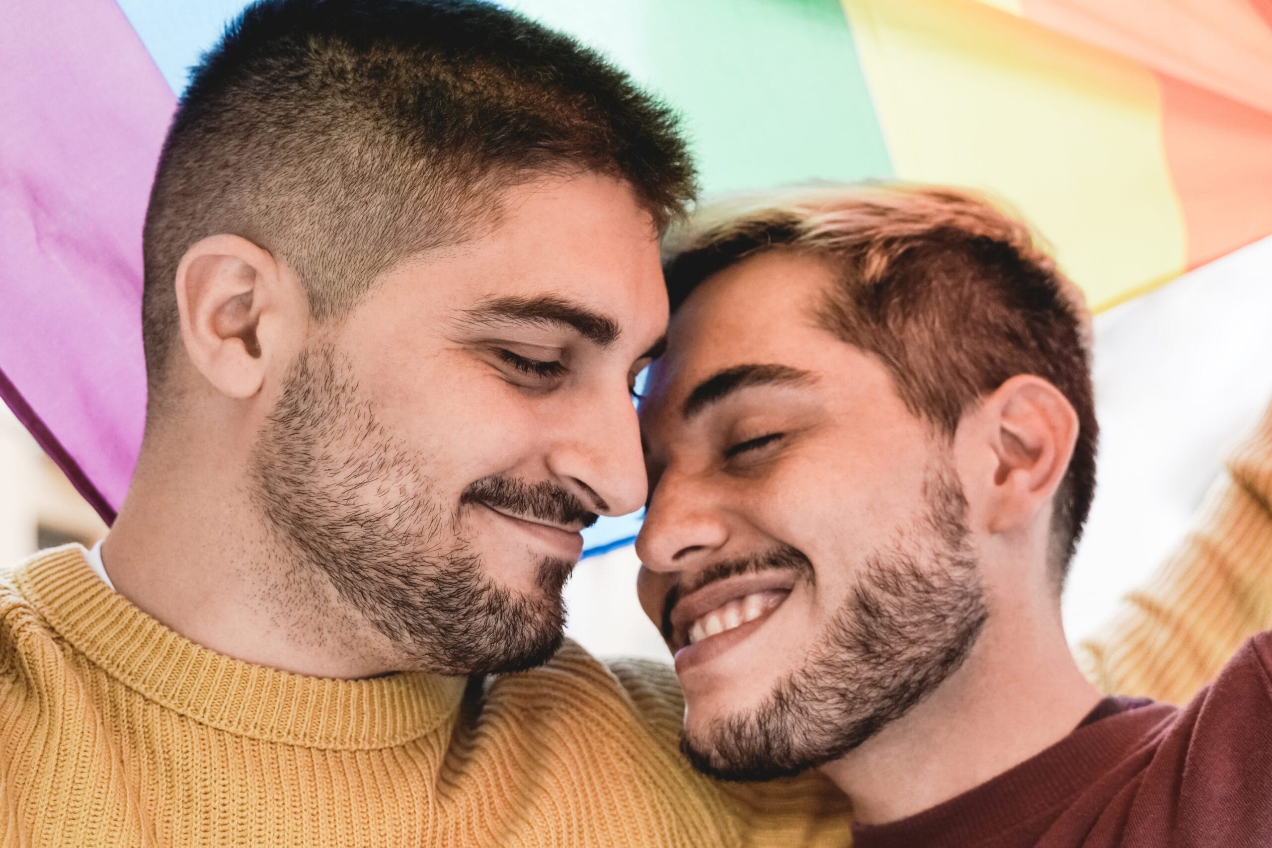 A tender moment between two smiling men with their foreheads touching, standing under a rainbow flag. One wears a mustard yellow sweater, and the other wears a maroon shirt, both radiating joy and affection. The image celebrates love, pride, and connection.