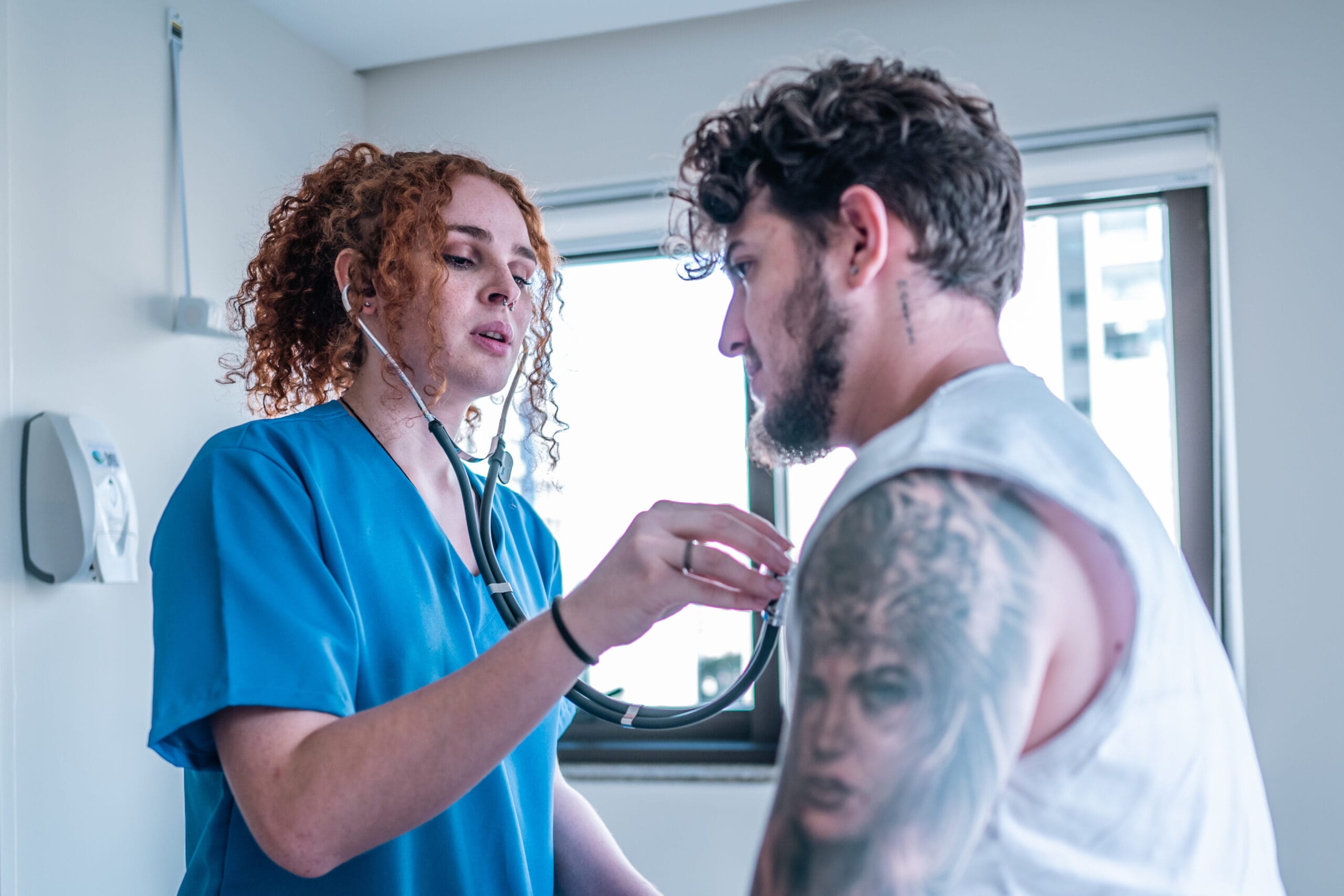 A healthcare professional with curly red hair in blue scrubs uses a stethoscope to examine a patient with tattoos on their arm in a clinical setting. The patient, wearing a sleeveless shirt, sits calmly as the provider focuses on the assessment. The room is well-lit with natural light coming through the windows.