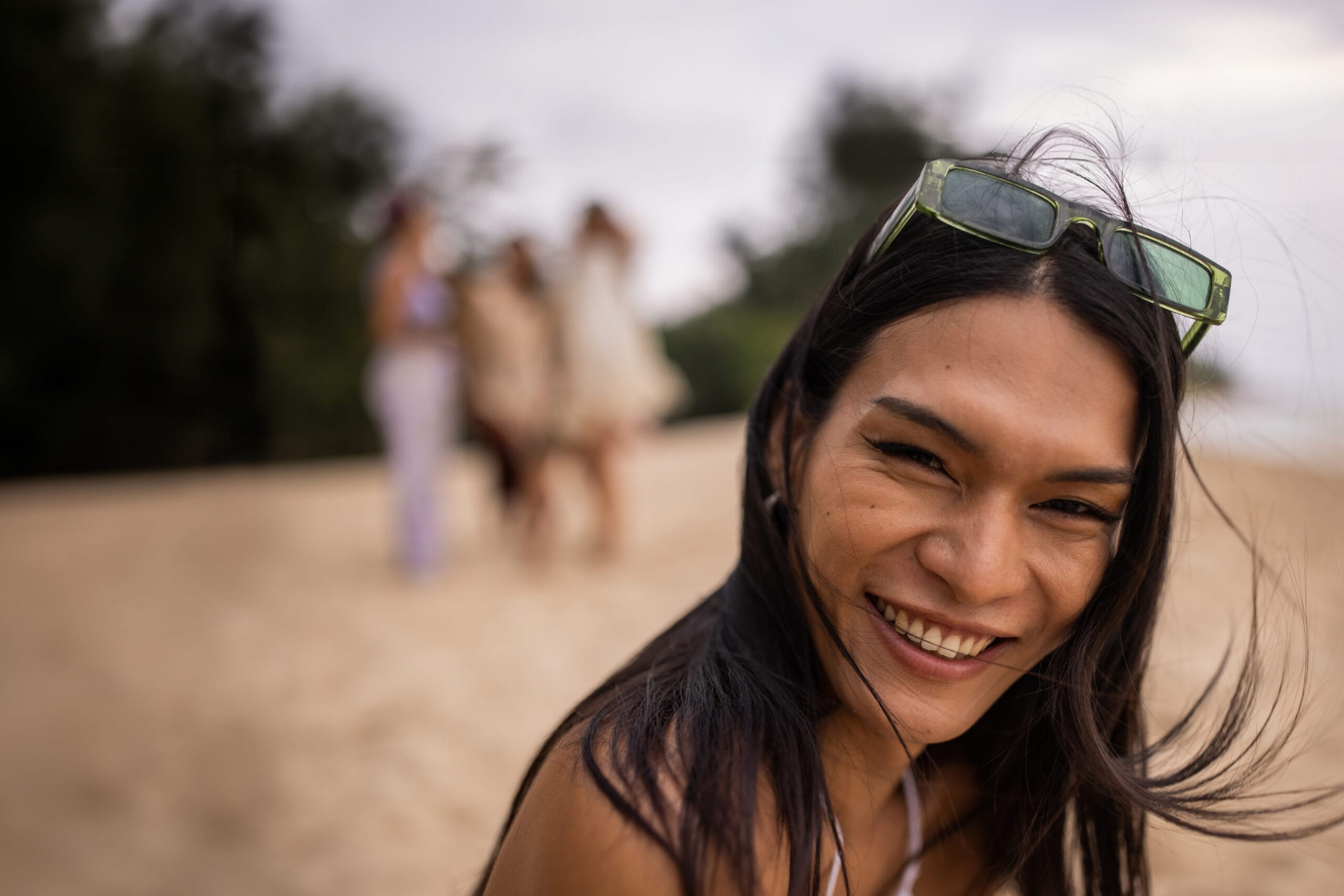 A joyful woman with long hair and green sunglasses resting on her head smiles brightly at the camera on a sandy beach. In the blurred background, a group of friends enjoys the beach together, creating a lively and carefree atmosphere. The image captures the warmth and happiness of a day spent outdoors.