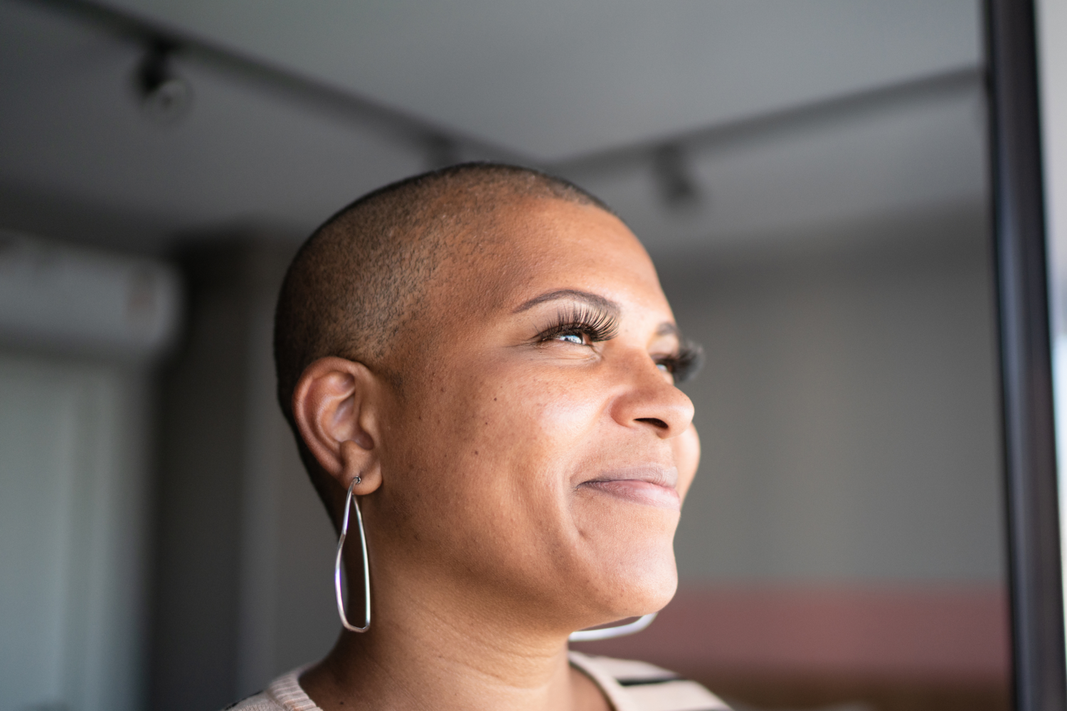 A close-up of a person with a shaved head and radiant skin, gazing out a window with a serene and confident expression. They wear long, teardrop-shaped earrings and natural makeup with long lashes, while soft natural light highlights their features. The setting conveys warmth and introspection.