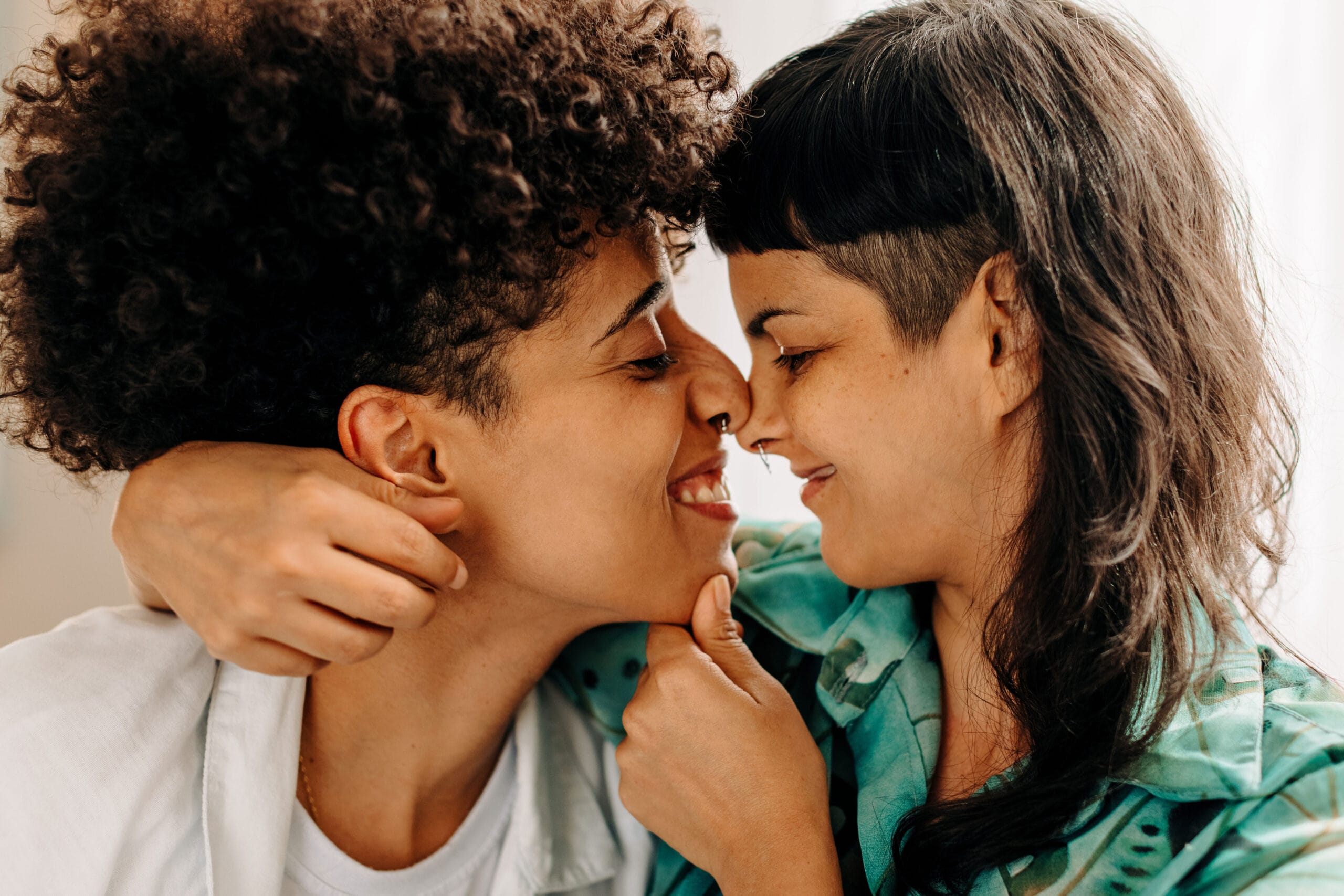 A tender moment between two people with noses touching and smiles on their faces, showcasing affection and connection. One has curly hair, and the other has a unique undercut hairstyle; both display septum piercings. The setting feels intimate and warm, highlighting love and closeness.