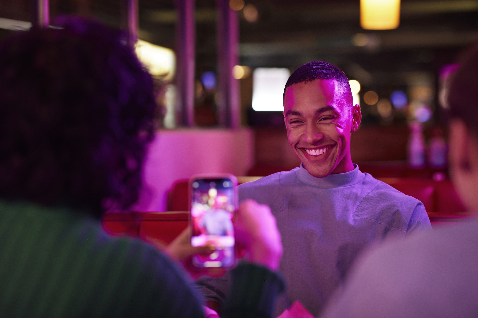 A cheerful person with a bright smile sitting in a warmly lit restaurant, illuminated by vibrant purple and yellow lights. The scene captures a casual moment as someone takes a photo of them with a smartphone, surrounded by friends in a cozy booth setting. The atmosphere feels lively and relaxed.