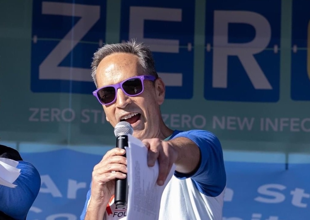 Glen Spencer wearing purple sunglasses and a blue and white shirt speaks energetically into a microphone while pointing towards the audience. Behind him is a banner with the word "ZERO" prominently displayed, along with phrases promoting health initiatives like "Zero Stigma" and "Zero New Infections." His dynamic expression and posture convey enthusiasm and engagement.