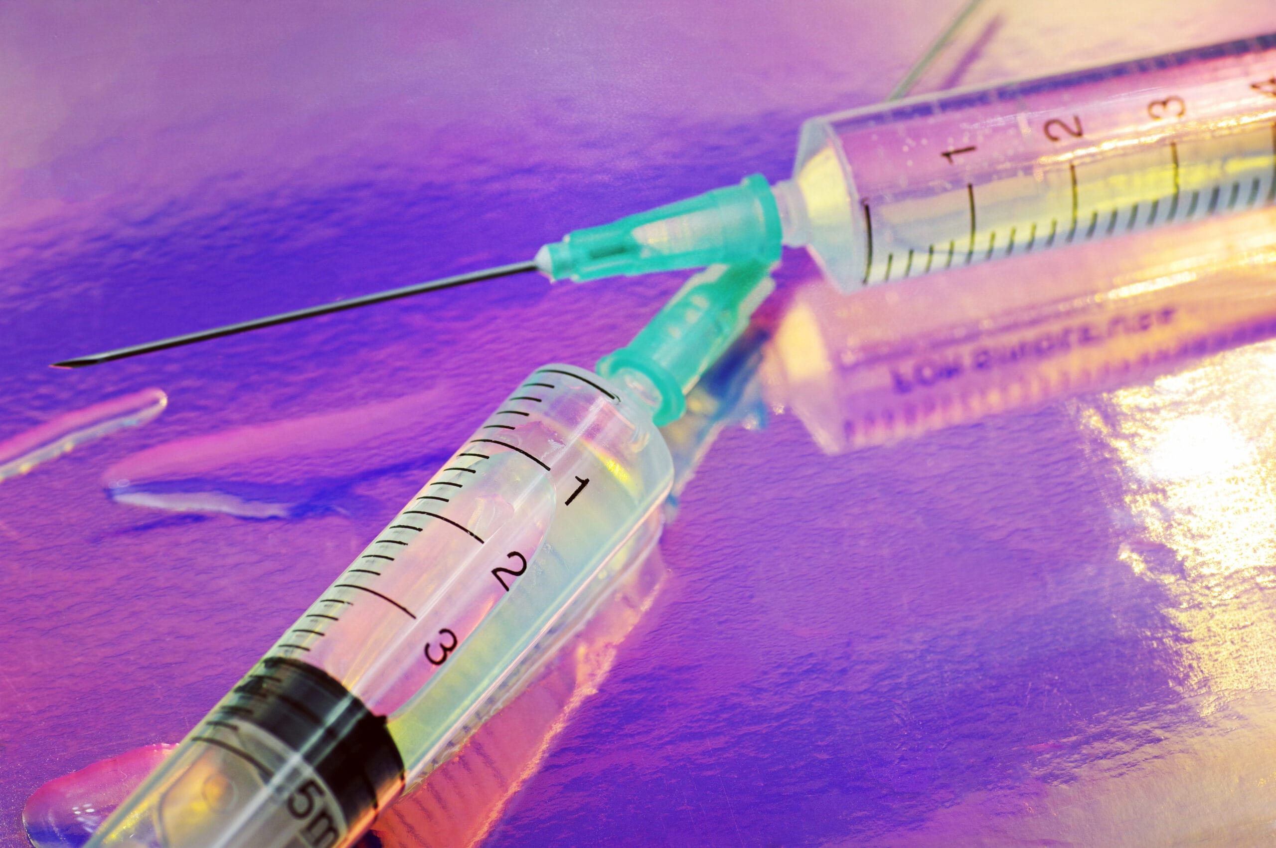 Two medical syringes filled with liquid are placed on a reflective surface with vibrant purple and pink lighting, creating a striking, futuristic aesthetic. The markings on the syringes and the needle tips are clearly visible, emphasizing a medical or scientific context. The colorful background adds a creative and eye-catching element to the composition.