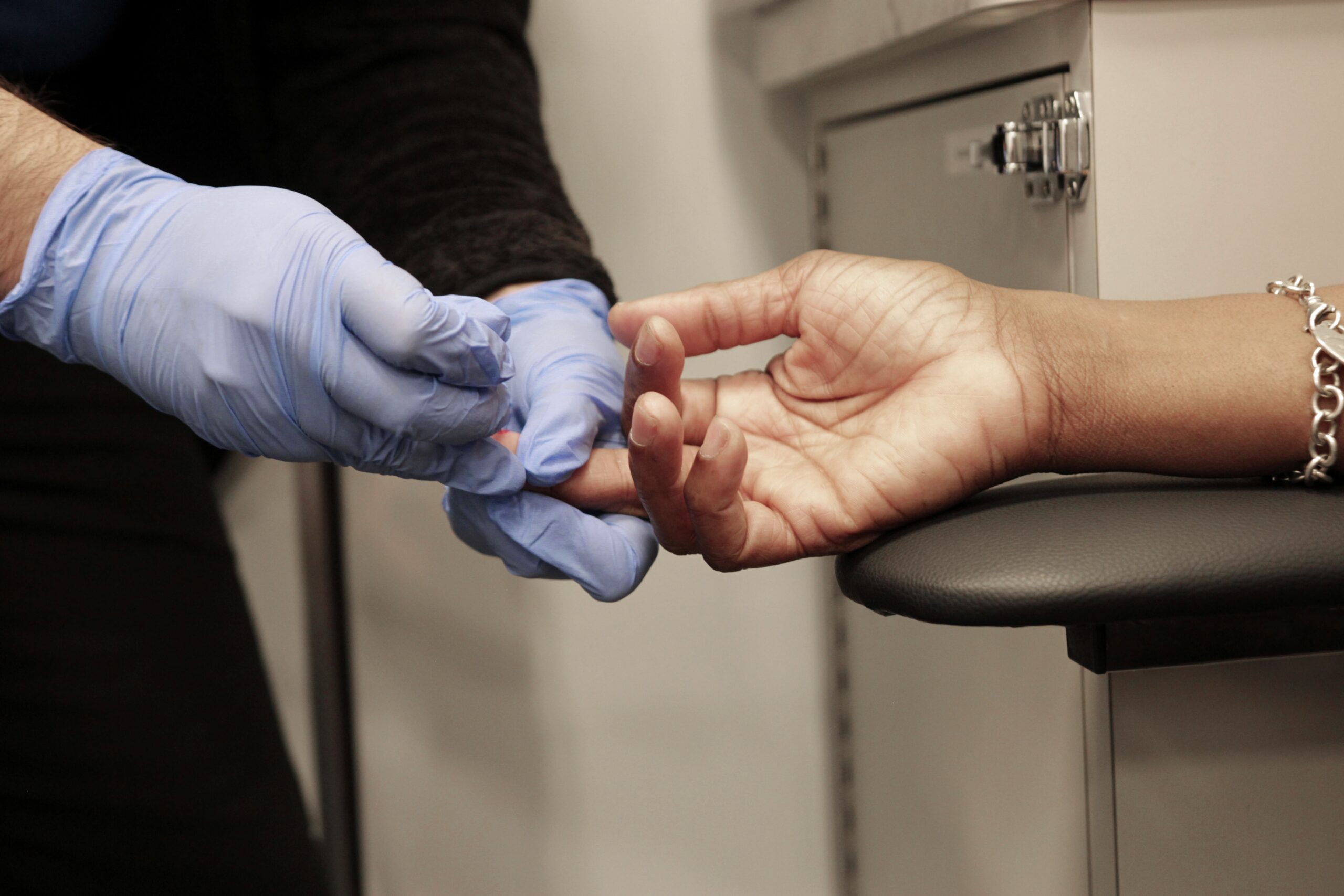 A person wearing blue gloves pricking a patient's finger with a lancet for a blood test, likely for a routine medical check-up or to monitor a health condition.