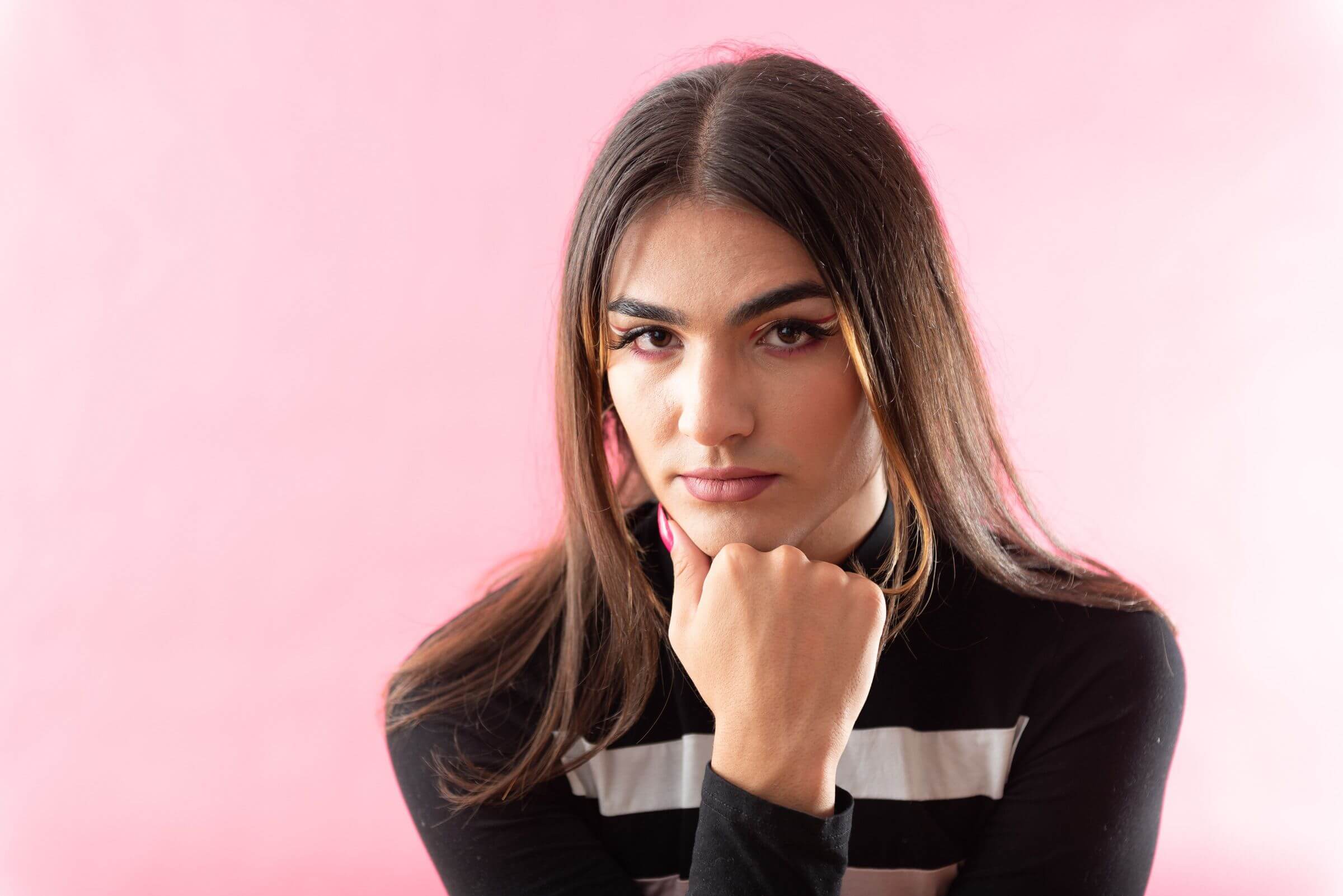 A person with long, sleek hair and sharp eyeliner gazes confidently at the camera, resting their chin on their hand. They wear a black and white striped top, and the soft pink background enhances the clean and polished composition. The overall mood is poised and empowering.