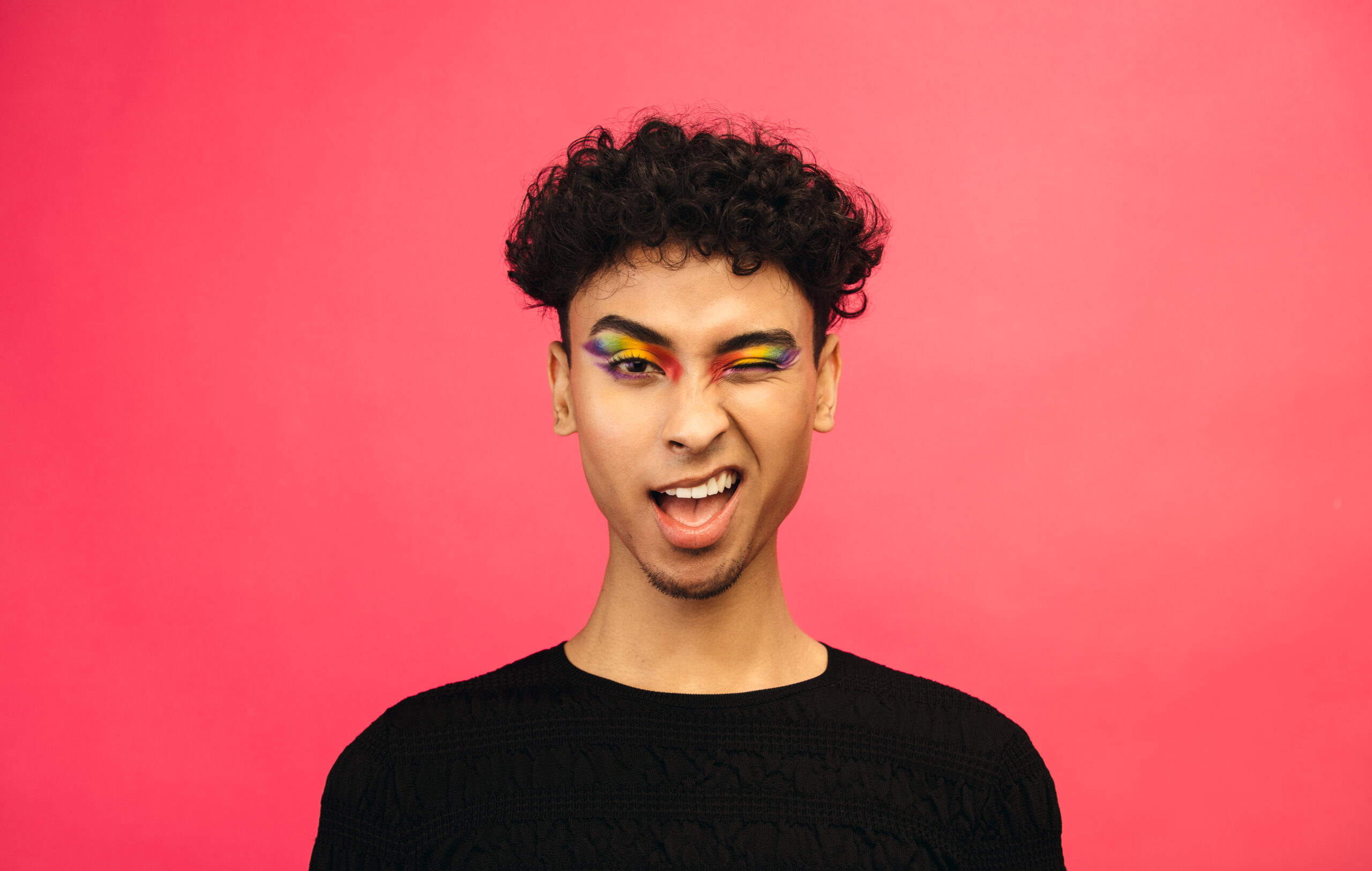 A person with curly hair wearing vibrant rainbow-colored eyeshadow, winking and smiling playfully against a bright pink background. They are dressed in a black textured top, exuding confidence and creativity.