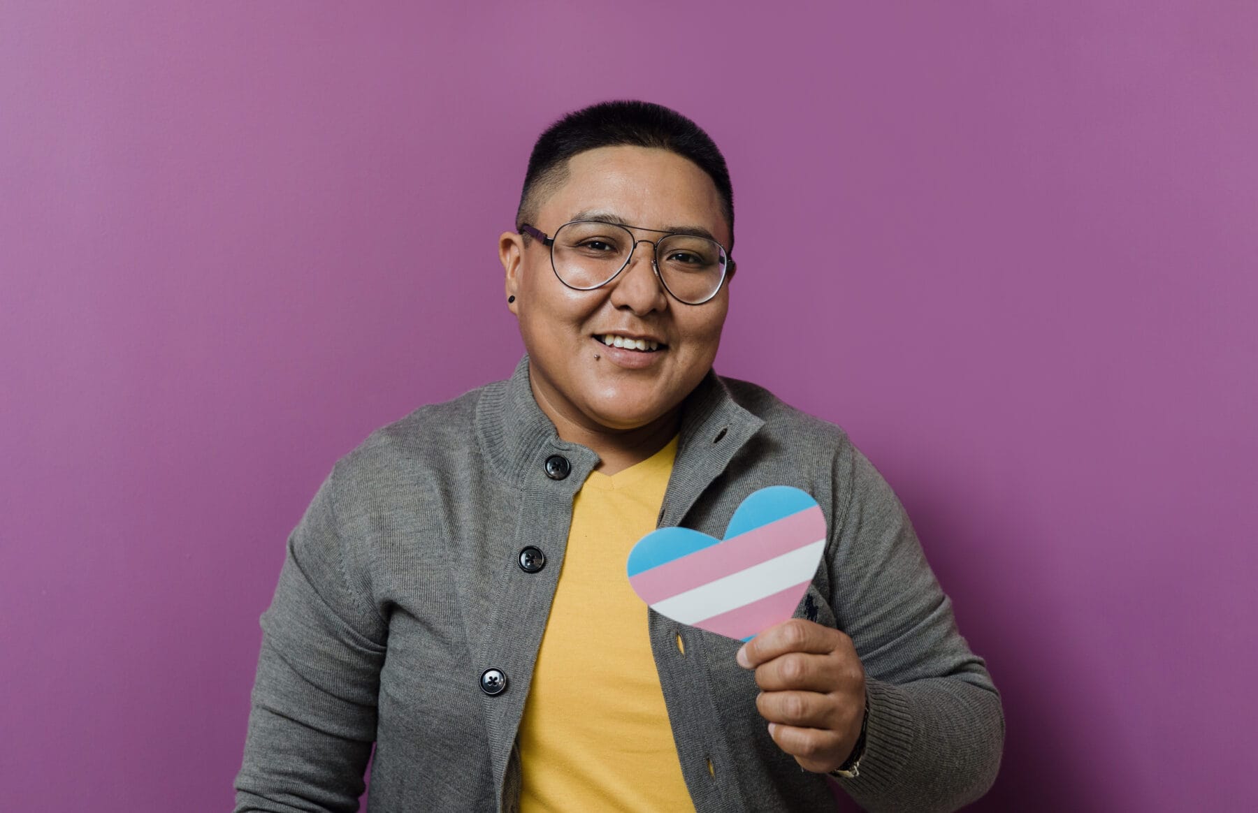 A smiling person wearing glasses and a gray cardigan over a yellow shirt, holding a heart-shaped cutout with the transgender pride flag design. They are standing against a vibrant purple background, radiating warmth and inclusivity.