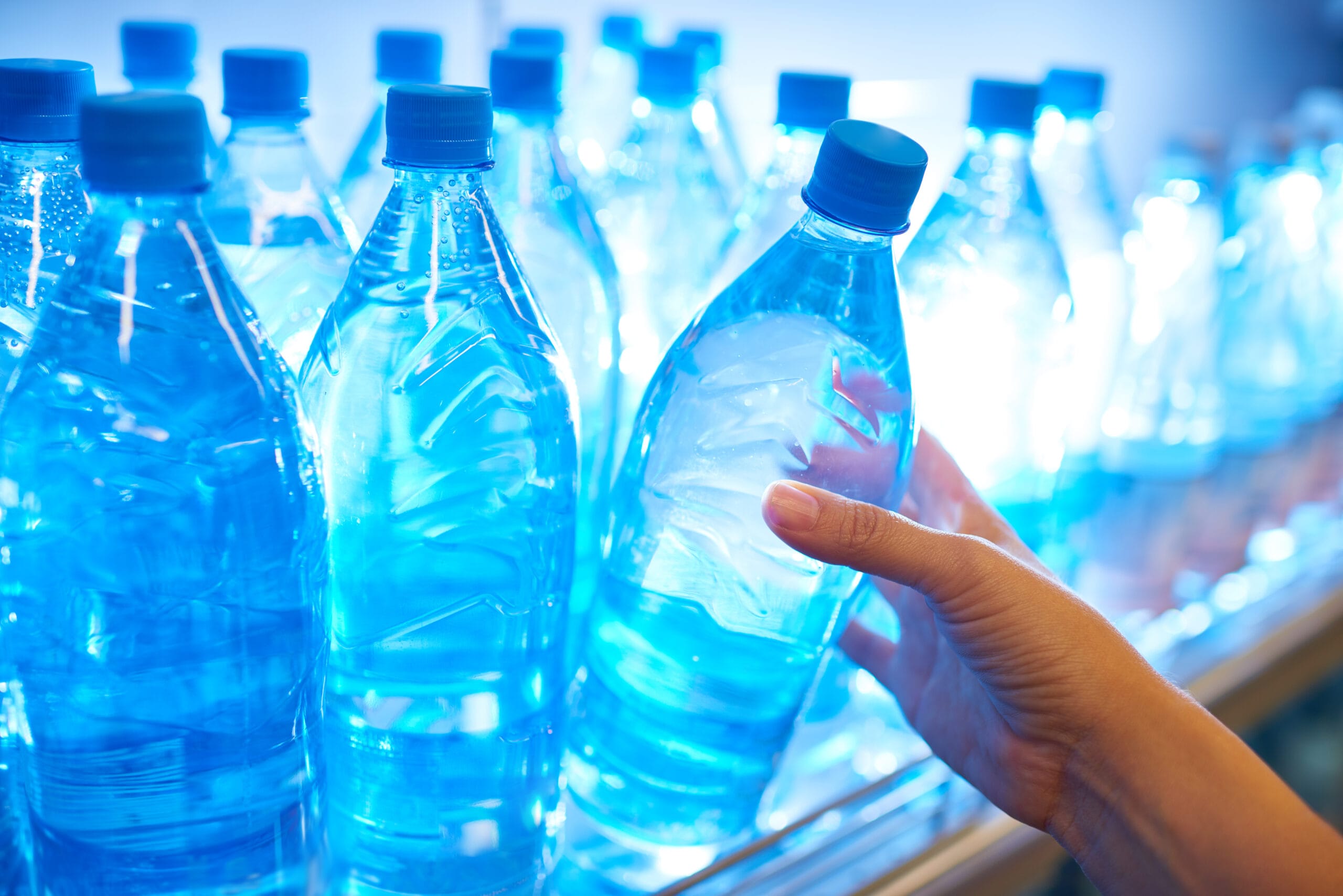 A row of clear plastic water bottles with blue caps is illuminated by bright lighting, creating a cool and refreshing ambiance. A hand reaches out to pick one bottle, emphasizing interaction and choice. The scene reflects hydration and convenience, with the light enhancing the clarity of the water inside the bottles.