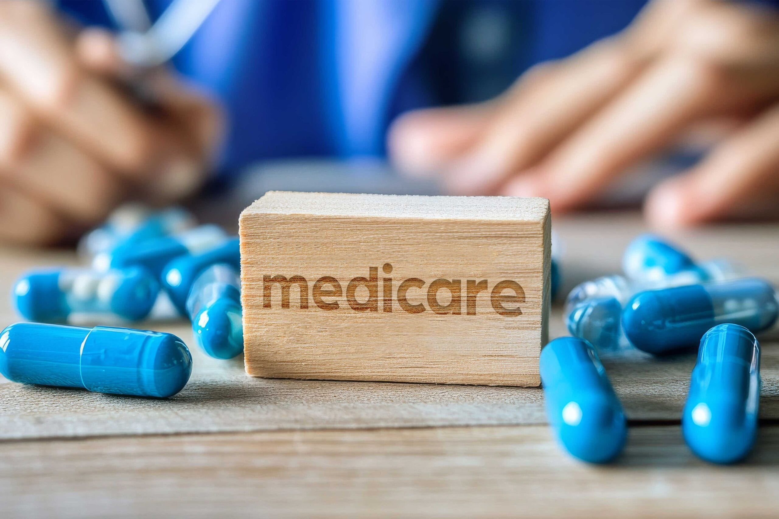 A wooden block with the word "Medicare" engraved on it, resting on a table with blue pills scattered around. A person in a blue medical uniform is blurred in the background, likely a doctor or healthcare professional. This image represents the concept of Medicare coverage for prescription medications.