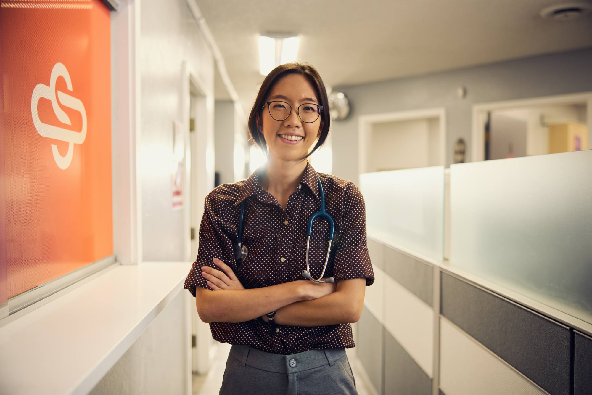 A smiling healthcare professional standing confidently in a brightly lit hallway, wearing a brown polka-dot shirt and a stethoscope around their neck. The environment is clean and modern, with an orange Spectrum Medical logo visible on a window in the background.