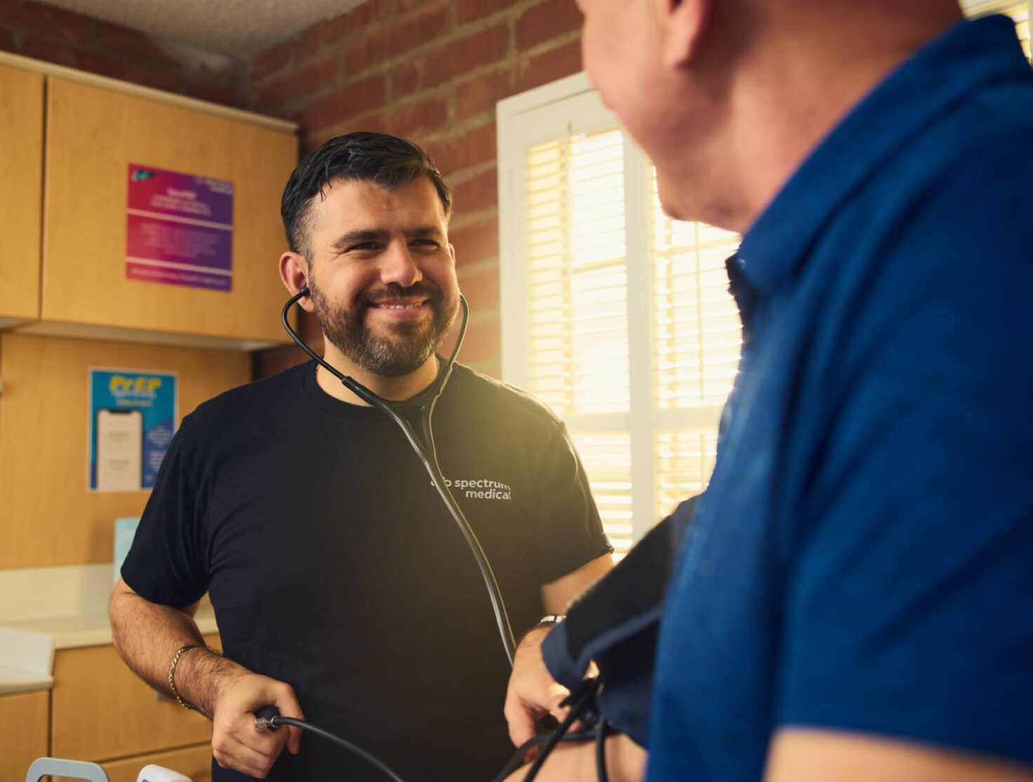 A healthcare professional wearing a "Spectrum Medical" shirt smiles warmly while using a stethoscope to check a patient's blood pressure. The setting is a sunlit clinic room with a brick wall and informational posters in the background. The interaction exudes a sense of care, trust, and professionalism.