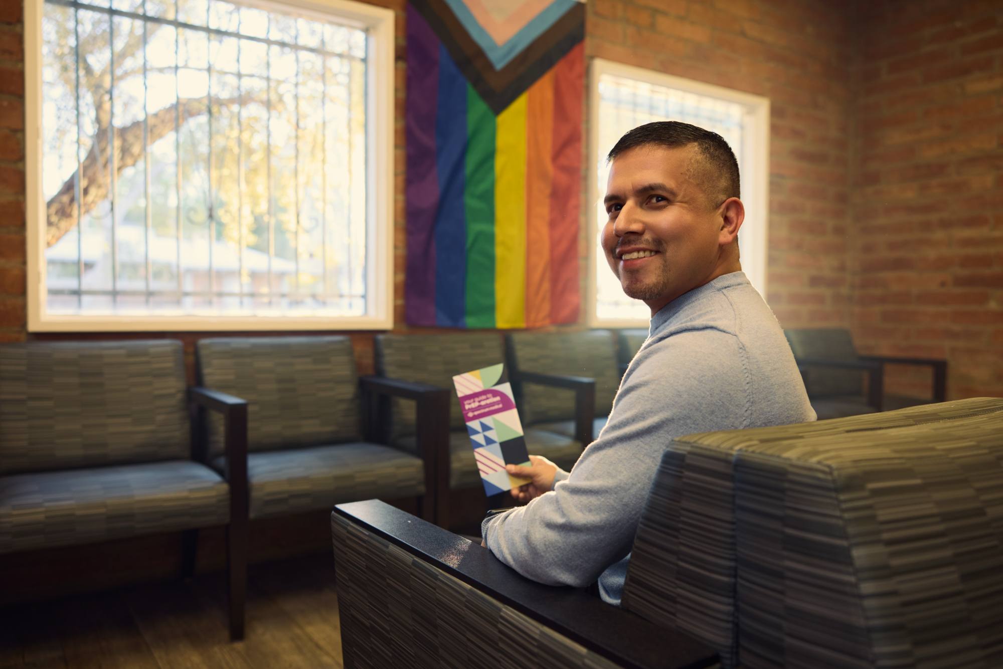 A man sits in a welcoming waiting area with modern chairs and a large window, holding an informational pamphlet. Behind him, a vibrant LGBTQIA2S+ Pride flag hangs on the brick wall, adding warmth and inclusivity to the space. He smiles toward the camera, creating a friendly and inviting atmosphere.