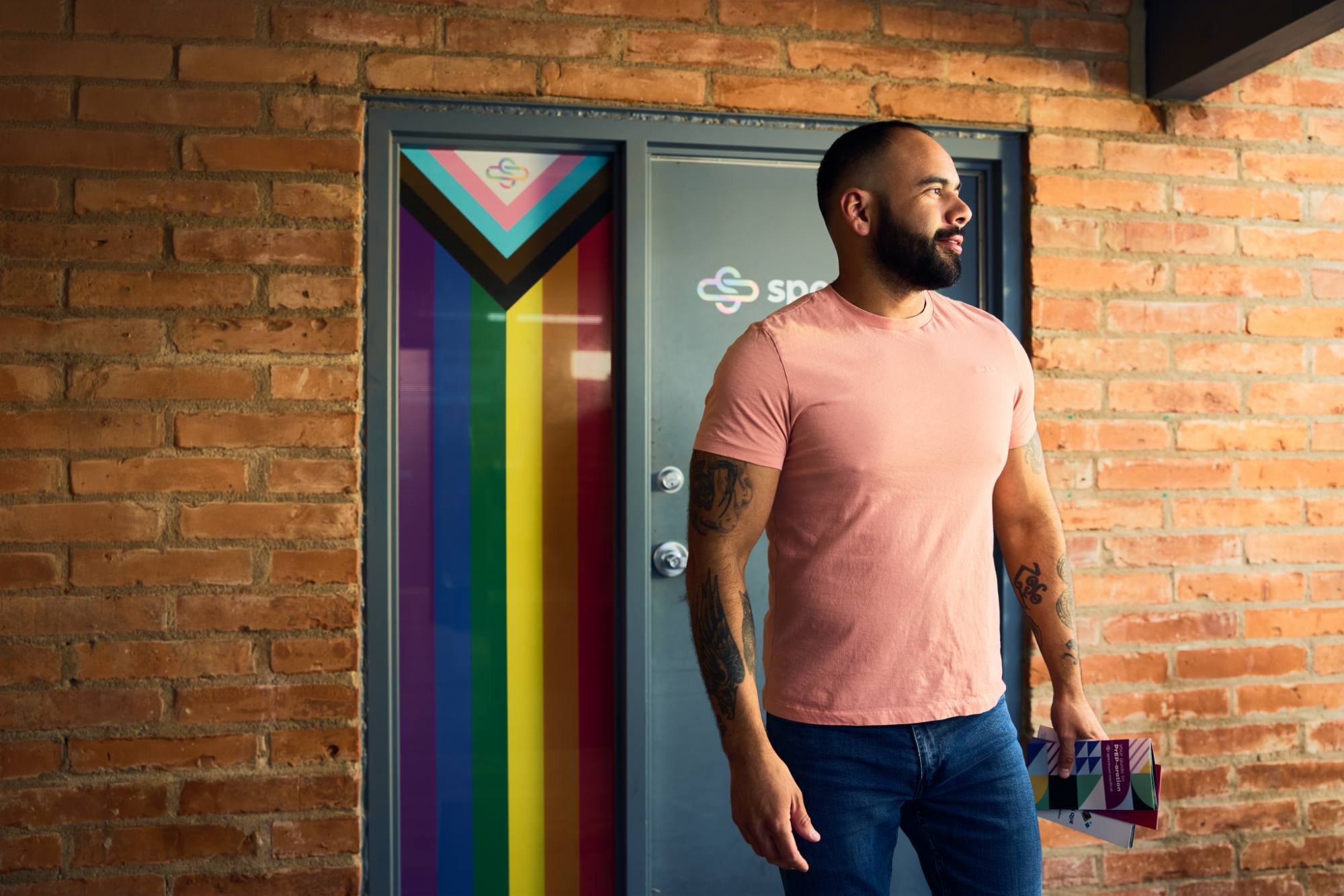 A man in a pink T-shirt and jeans stands in front of a brick wall, holding informational brochures. Behind him, a door is adorned with the Progress Pride flag design, symbolizing inclusivity and support.