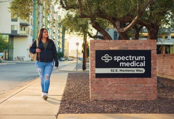A woman walks along a sidewalk near a brick sign for "Spectrum Medical, 52 E. Monterey Way." She wears a casual outfit with a backpack, set against a backdrop of urban buildings and trees in the warm glow of the evening light.