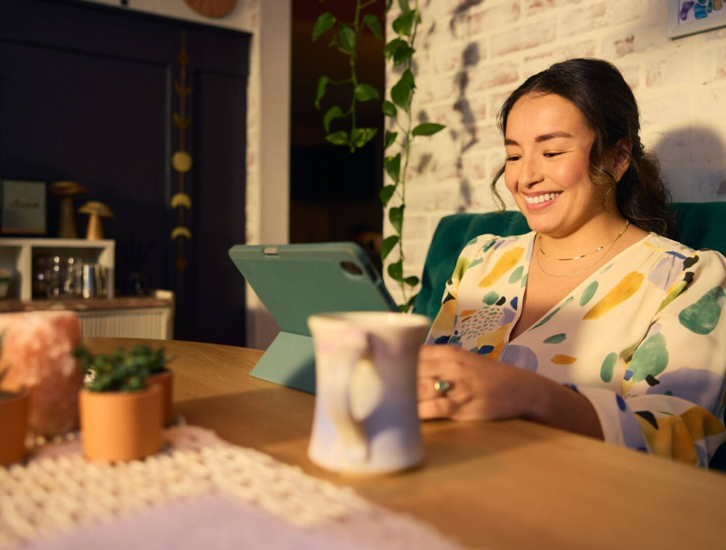 A smiling woman sits at a wooden table, engaged with a tablet in a cozy, well-decorated space. She wears a colorful patterned blouse and is surrounded by small potted plants, a mug, and soft lighting that enhances the warm and inviting atmosphere. The scene conveys comfort and focused relaxation.