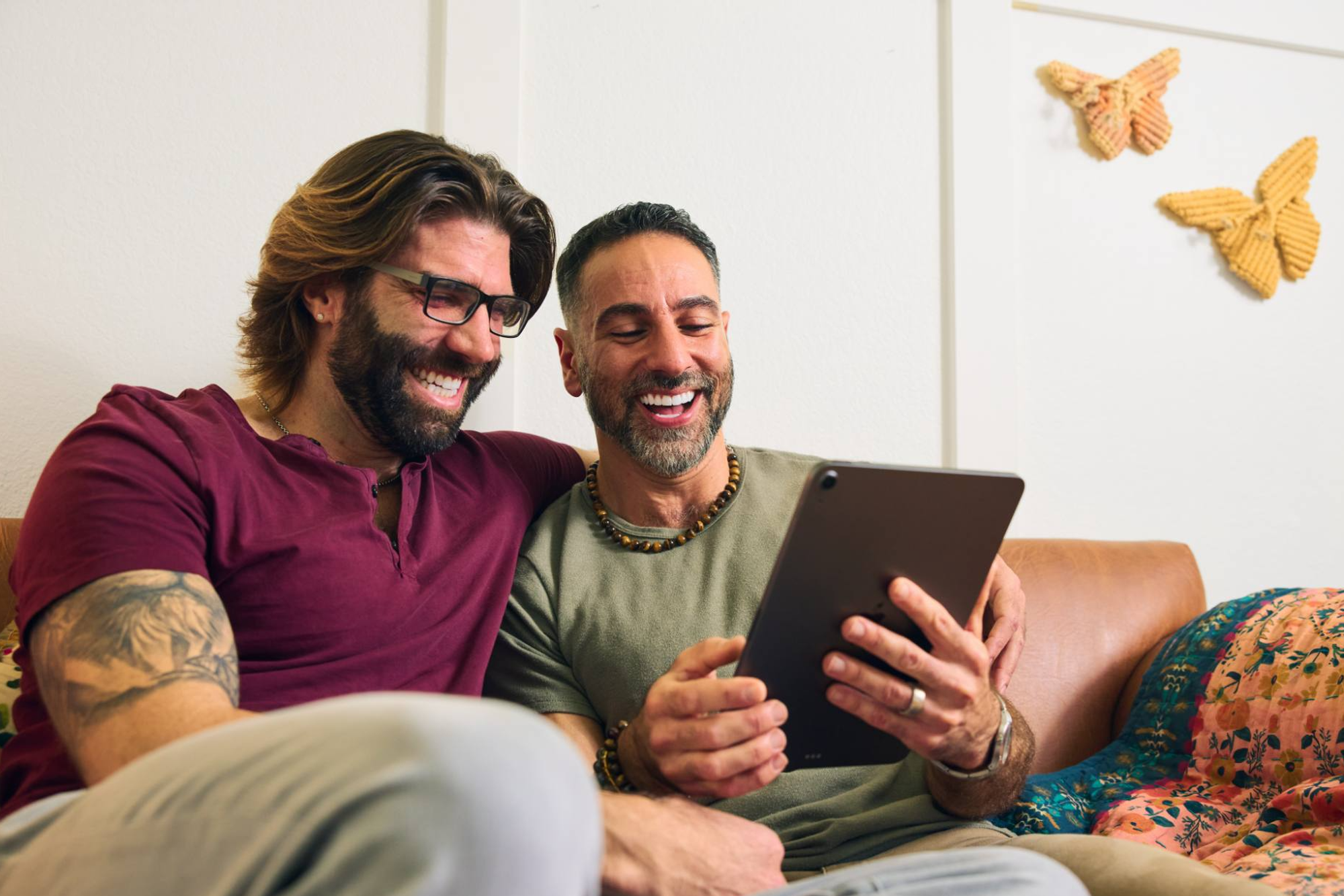 Two men sit closely on a couch, smiling and laughing as they look at a tablet screen together. One wears a maroon shirt with glasses, and the other sports a green shirt with a beaded necklace. The warm and inviting space is decorated with textured wall art and a colorful throw blanket.