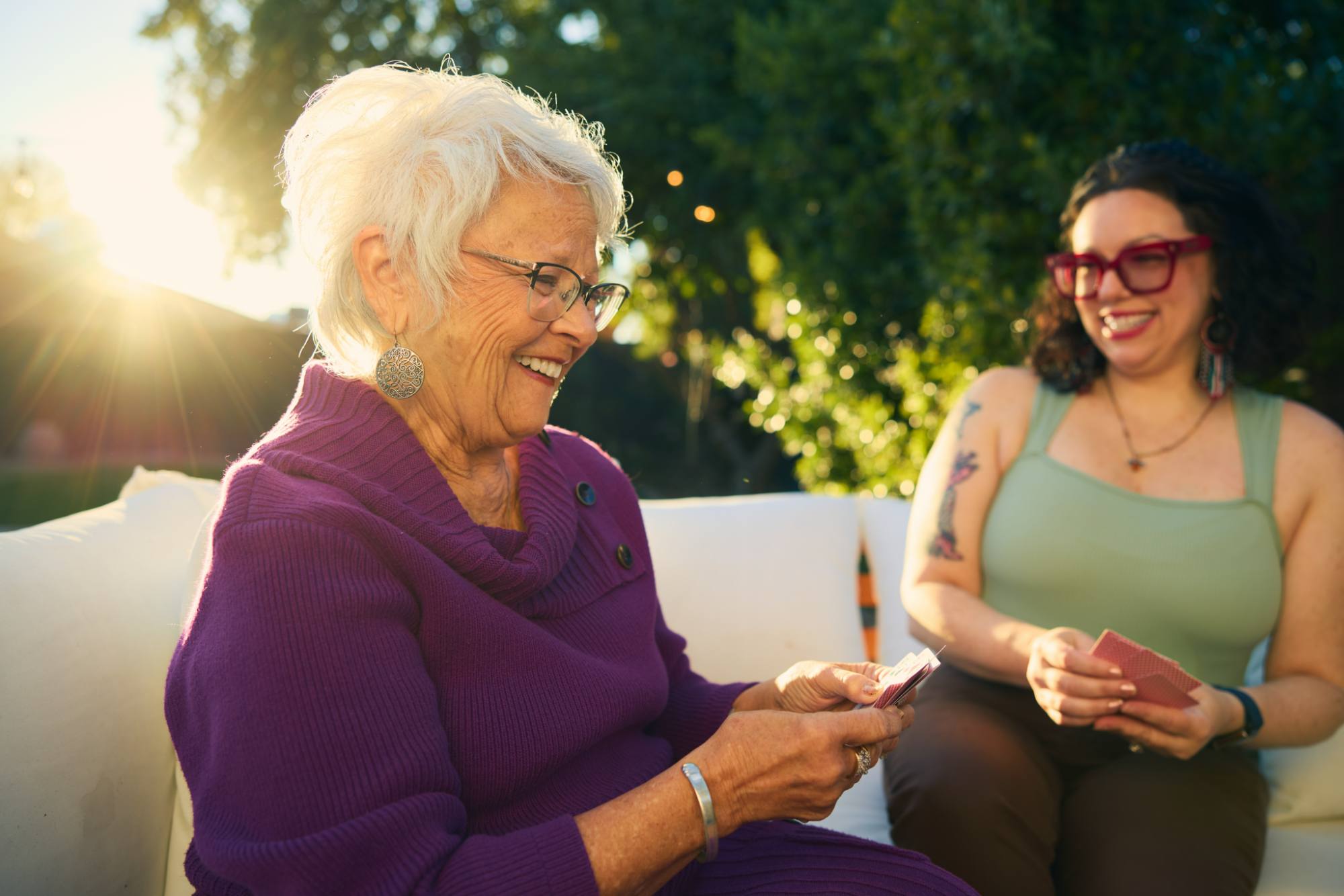 An elderly woman with white hair and glasses, dressed in a vibrant purple sweater, smiles warmly while playing cards with a younger woman sitting beside her. The scene is set outdoors in golden sunlight, with greenery in the background, evoking a joyful and relaxing atmosphere.