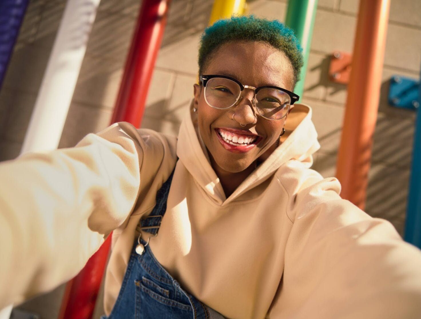 A cheerful Black person with short teal hair, wearing glasses, a beige hoodie, and denim overalls, takes a selfie against a colorful background of rainbow-colored vertical poles. They are smiling brightly, exuding joy and confidence in the warm sunlight. The image captures a lively and playful moment.