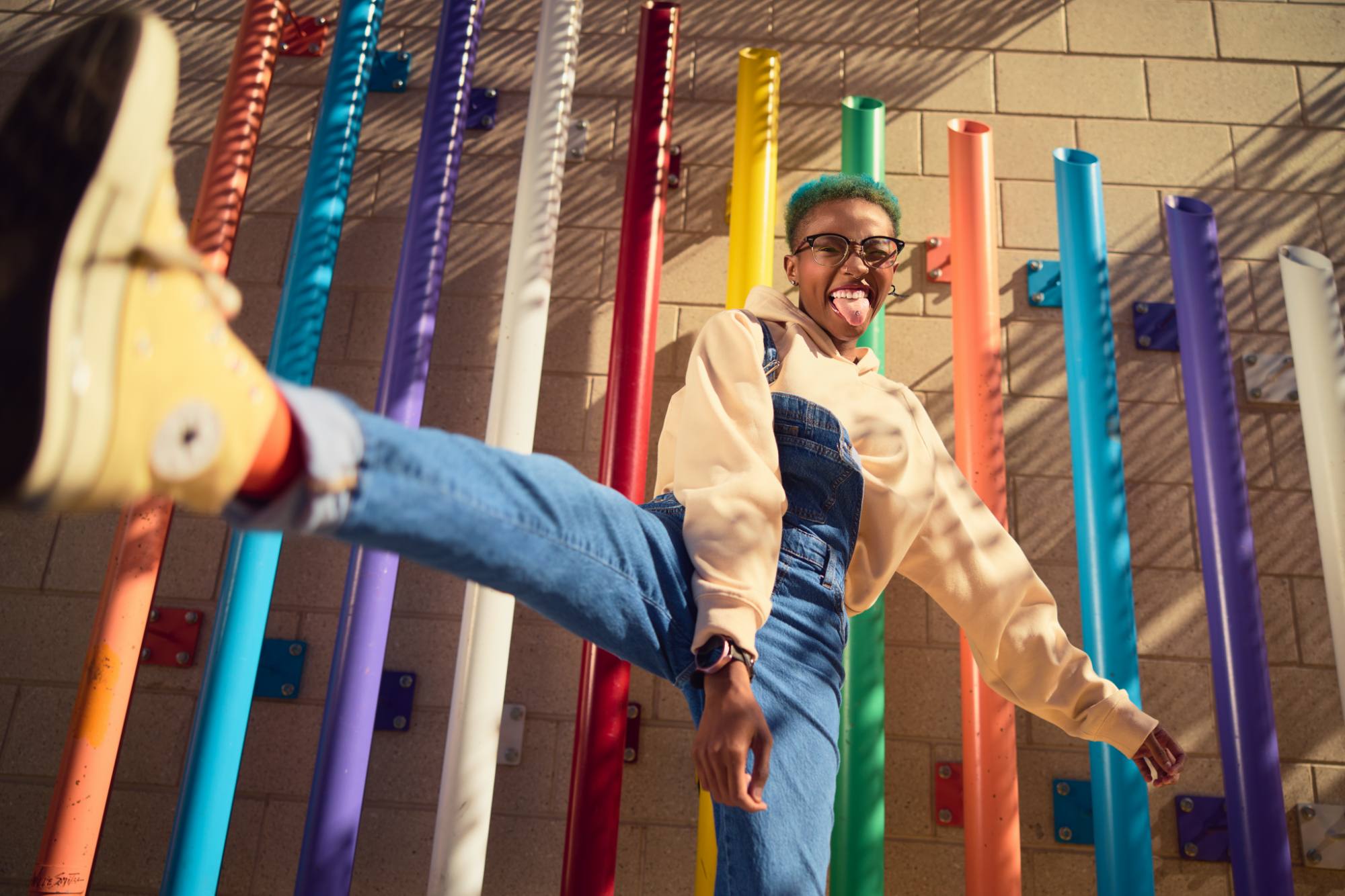 A playful Black person wearing overalls and a light-colored hoodie sticks out their tongue while kicking their foot playfully in front of a colorful wall of vertical pipes in various hues. The vibrant scene is filled with energy and fun.