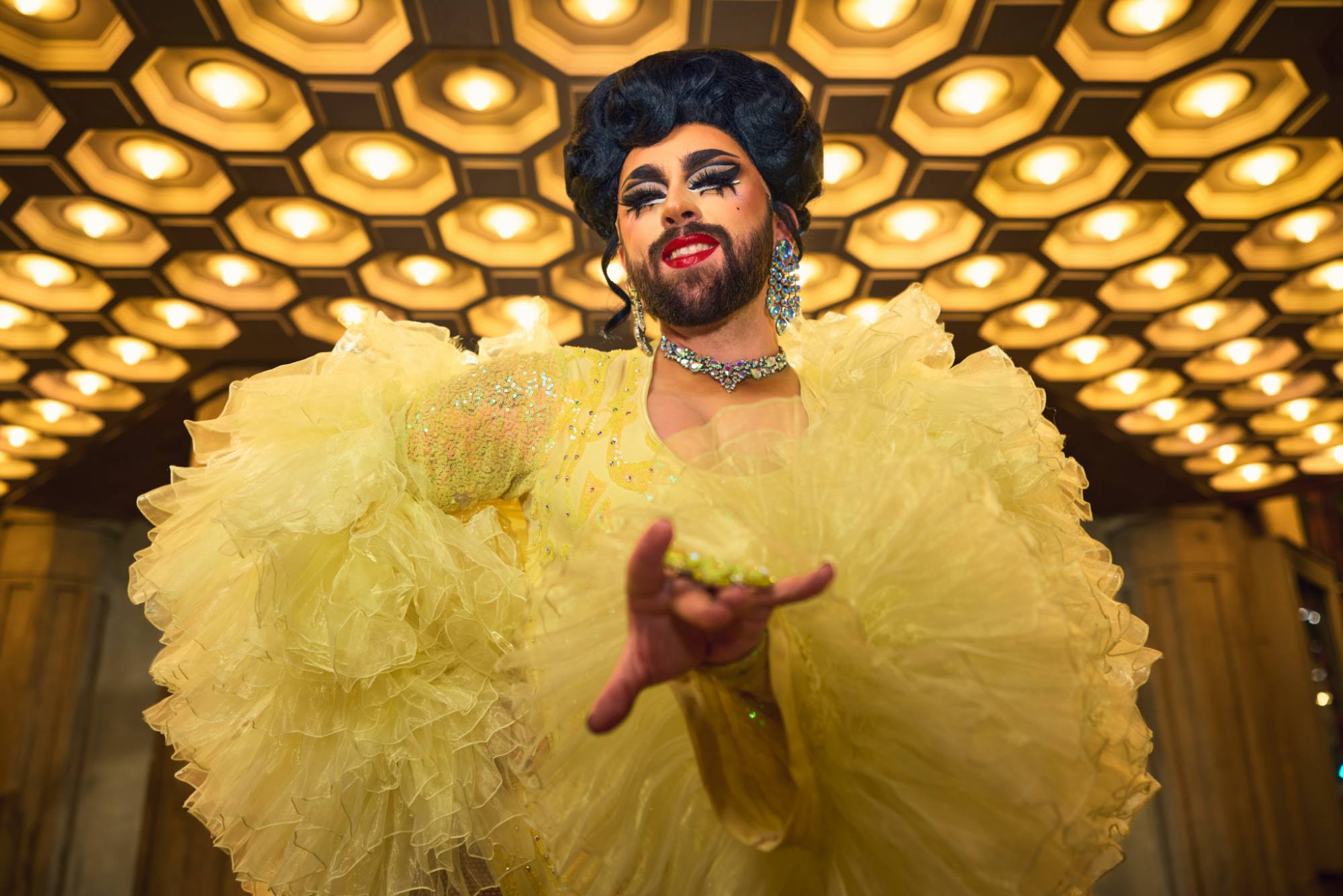 Trey DeGroodt - drag queen. A glamorous drag performer dressed in a voluminous, sparkling yellow gown poses confidently under a ceiling of glowing geometric lights. The performer exudes elegance and drama with bold makeup, intricate accessories, and a radiant smile.