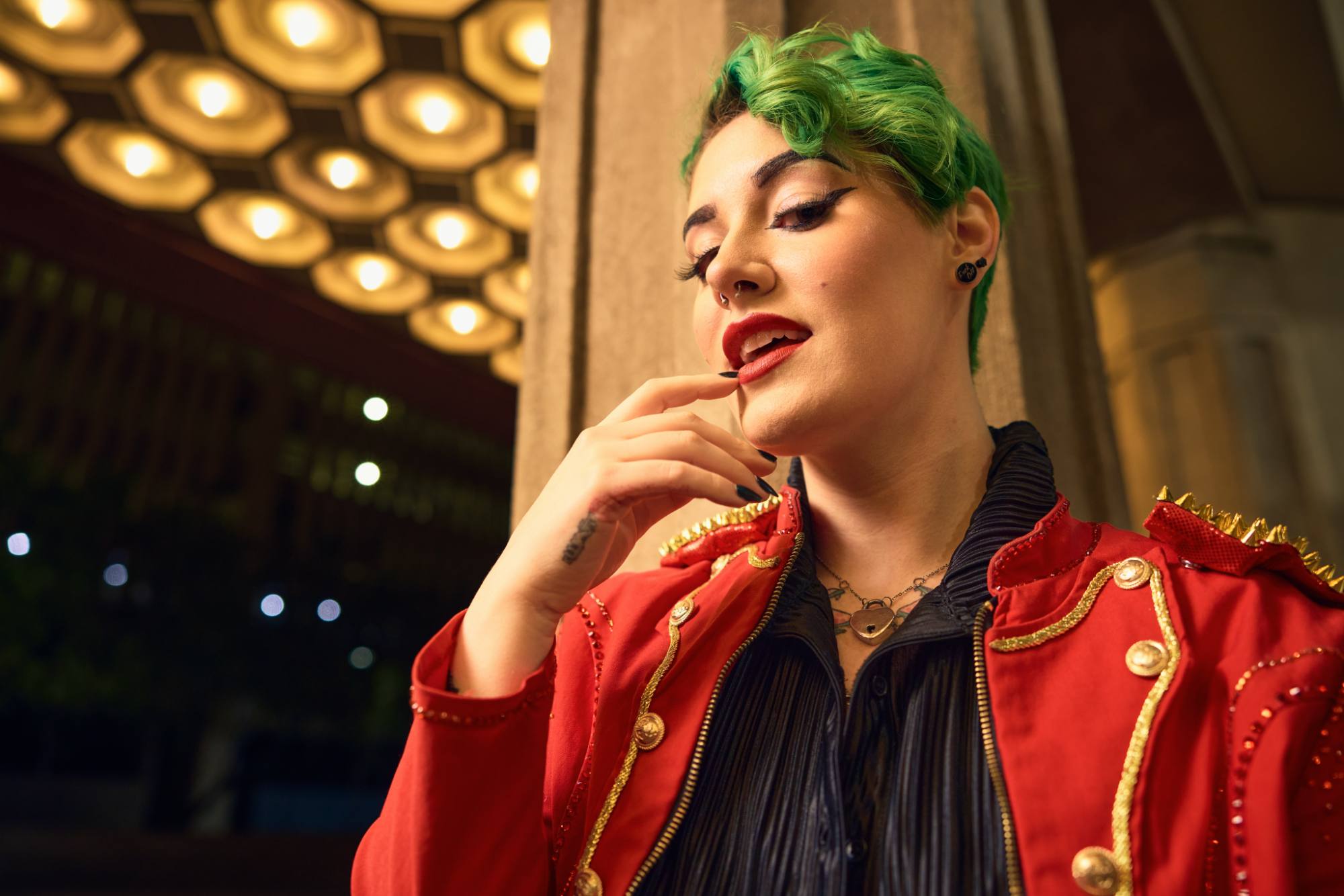 A person with vibrant green hair and bold makeup poses confidently under warm, decorative lighting. They wear a striking red jacket adorned with gold embellishments and spikes, paired with a black textured shirt, exuding a dramatic and stylish flair.