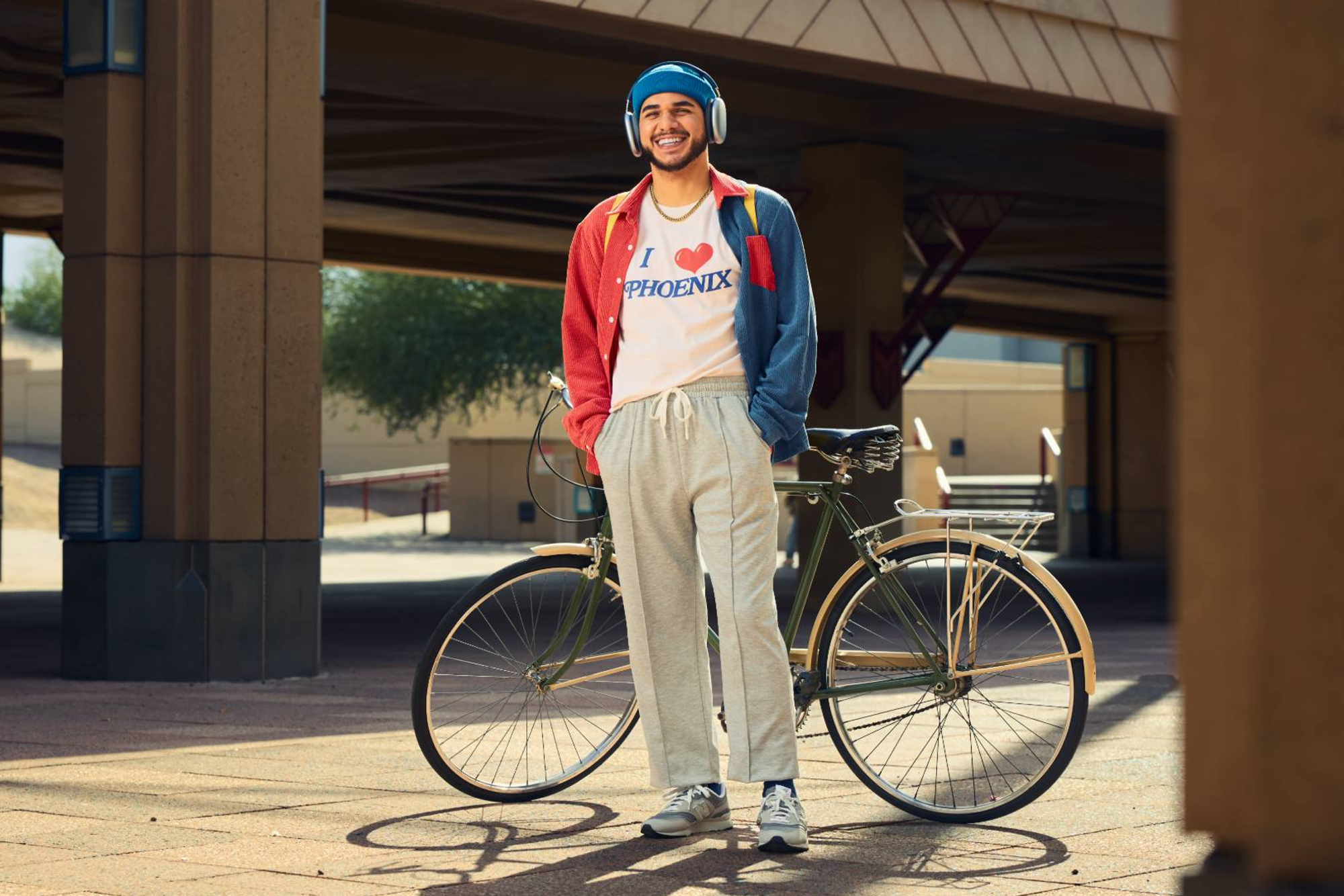 A young man stands confidently next to a vintage-style bicycle under a modern overpass. He wears headphones, a blue beanie, and a colorful jacket over a shirt that reads "I ❤️ Phoenix," paired with gray sweatpants and sneakers. The warm outdoor setting complements his relaxed, urban vibe.