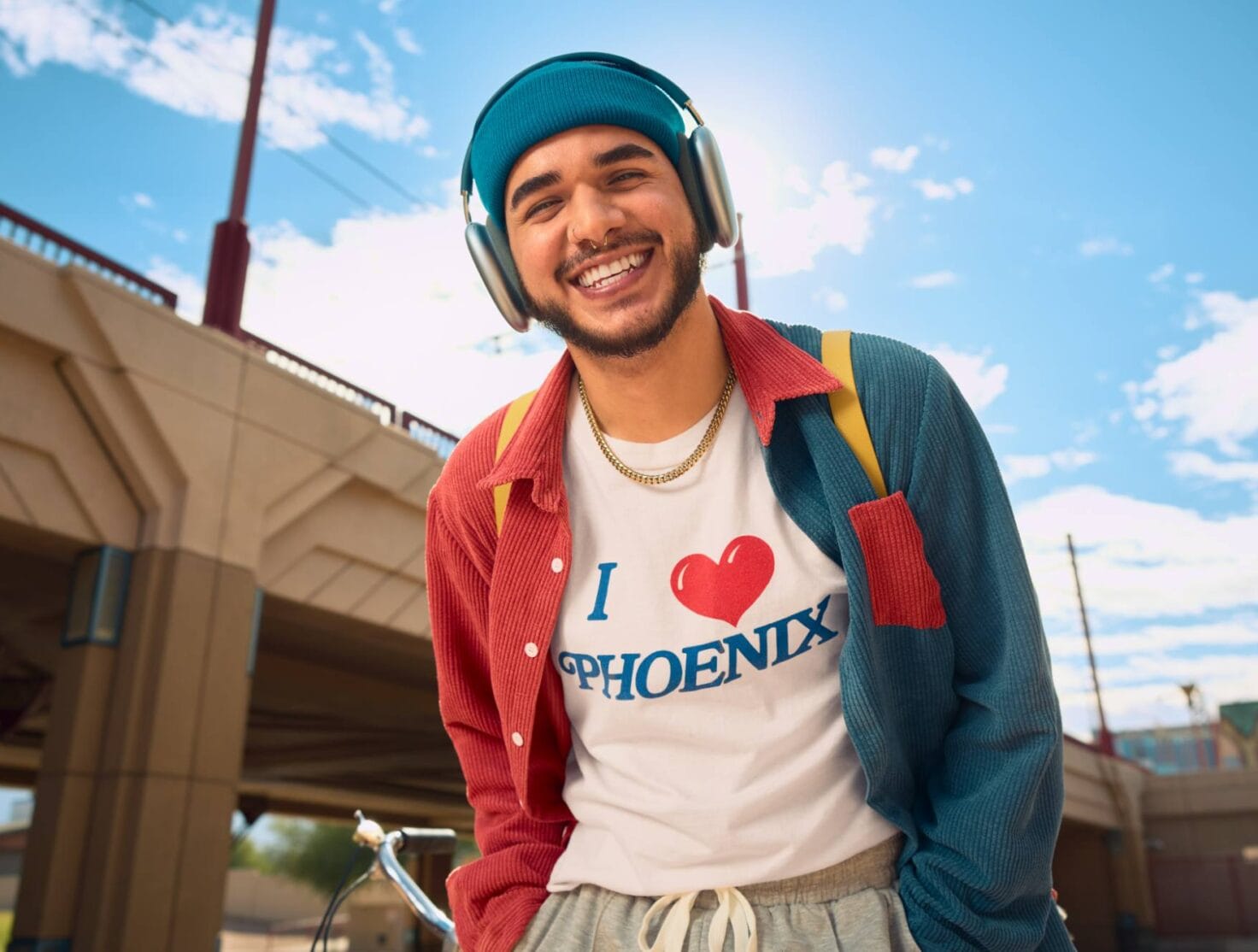 A young man smiles brightly outdoors, wearing a blue beanie, headphones, and a colorful red and teal shirt over a white T-shirt that reads "I ❤️ PHOENIX." He has a yellow backpack strap on his shoulder, with a sunny sky and an urban bridge structure in the background, capturing a vibrant and cheerful vibe.