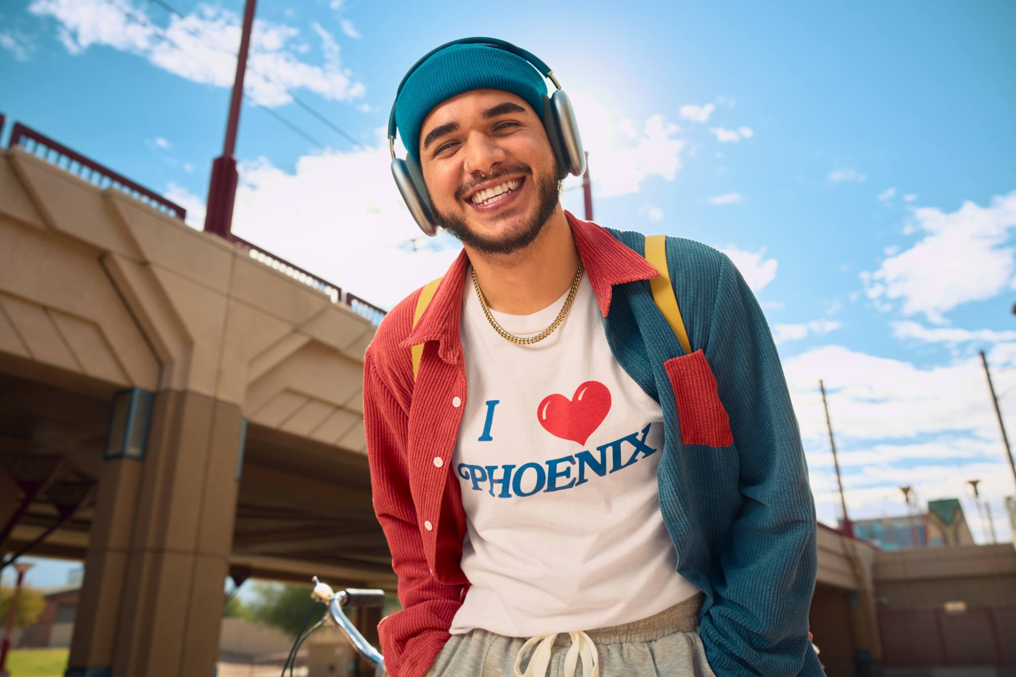 A young man smiles brightly outdoors, wearing a blue beanie, headphones, and a colorful red and teal shirt over a white T-shirt that reads "I ❤️ PHOENIX." He has a yellow backpack strap on his shoulder, with a sunny sky and an urban bridge structure in the background, capturing a vibrant and cheerful vibe.