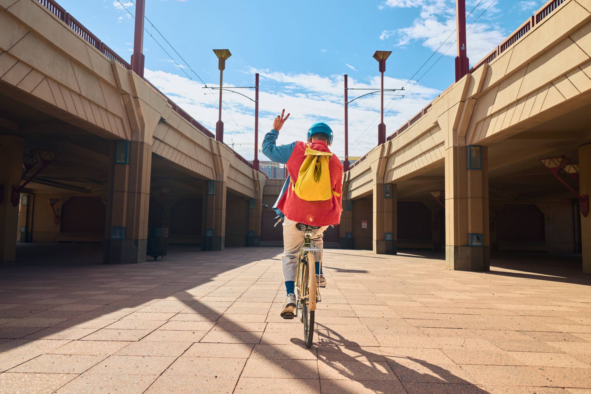 A cyclist rides through a parking facility on a sunny day, wearing a vibrant yellow backpack and a helmet, raising a hand in a peace sign. The modern architecture of the covered parking lot frames the scene, with a clear blue sky above.