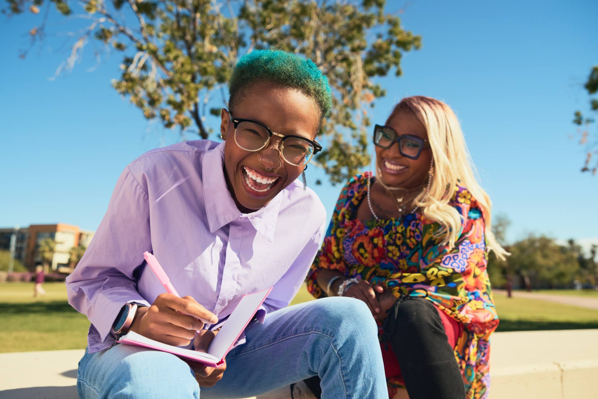 Two joyful individuals enjoying a sunny day outdoors, one with teal short hair and glasses writing in a notebook while laughing, and the other with long blond hair and bold glasses smiling warmly. The vibrant colors of their outfits and the bright blue sky create a lively and cheerful atmosphere, radiating friendship and positivity.